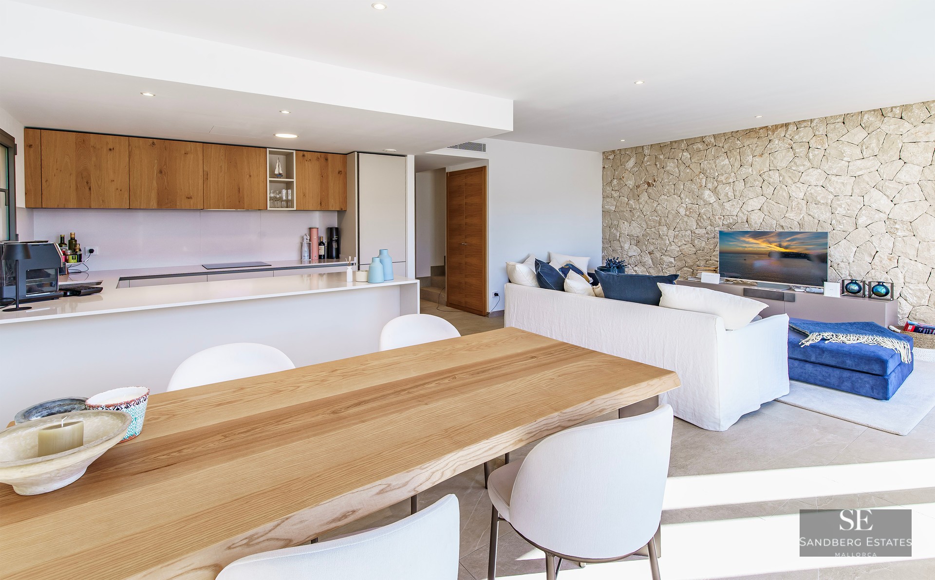 Bright open-plan room with wooden dining table, modern kitchen, white sofa, and a textured stone feature wall.
