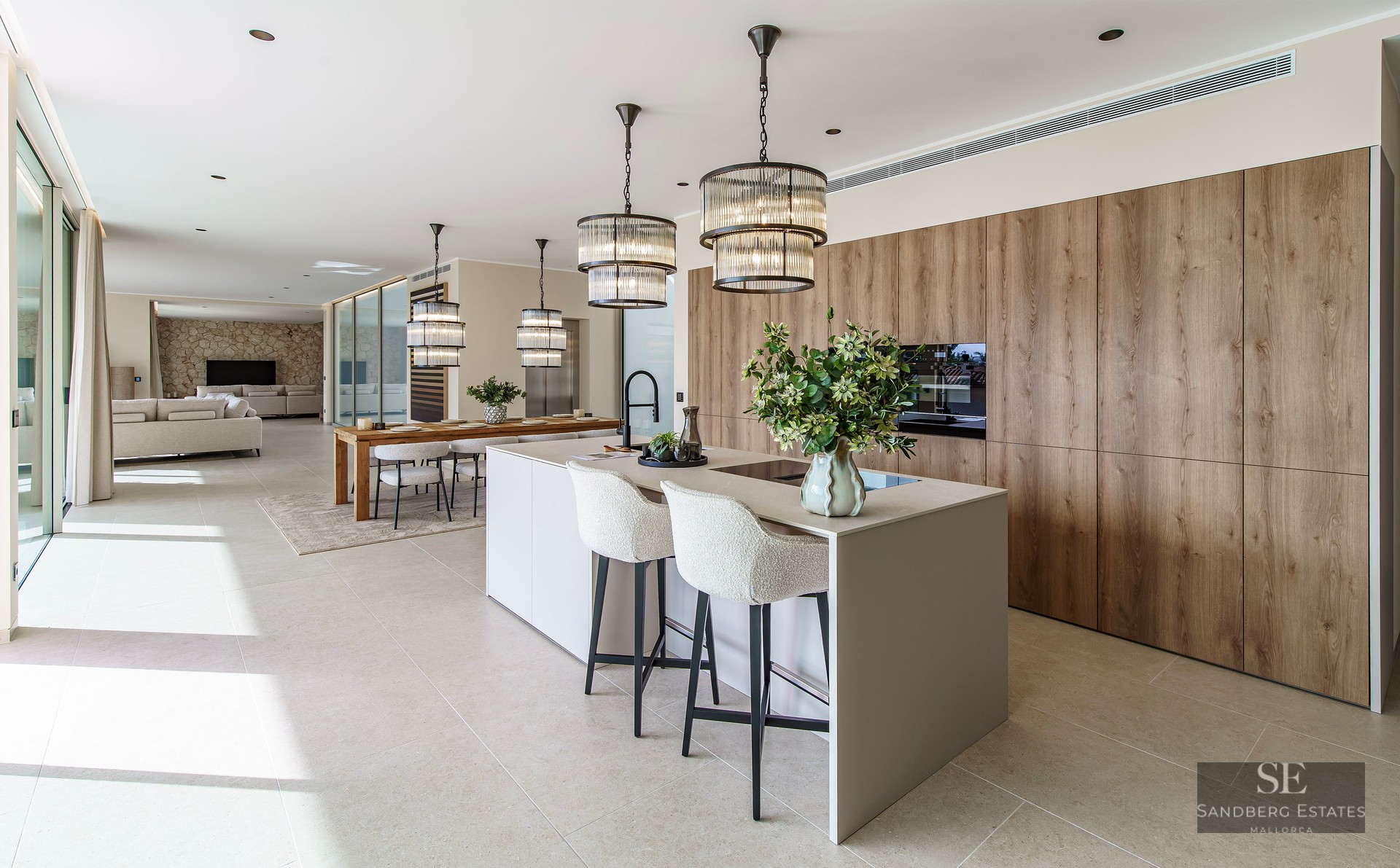Modern kitchen with a large island, white stools, designer pendant lights, and wood cabinetry in an open-plan space.