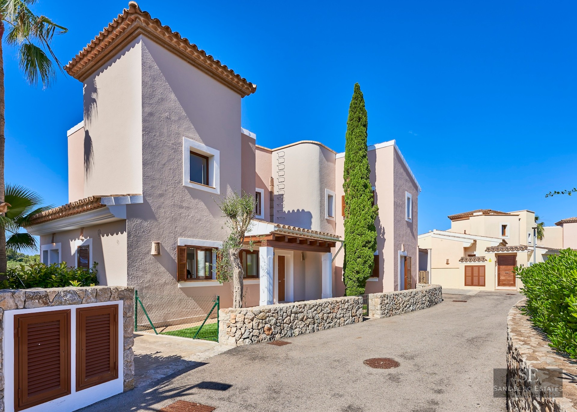 Mediterranean-style villa in pale terracotta with stone walls, wooden shutters, and a tall cypress tree under a blue sky.