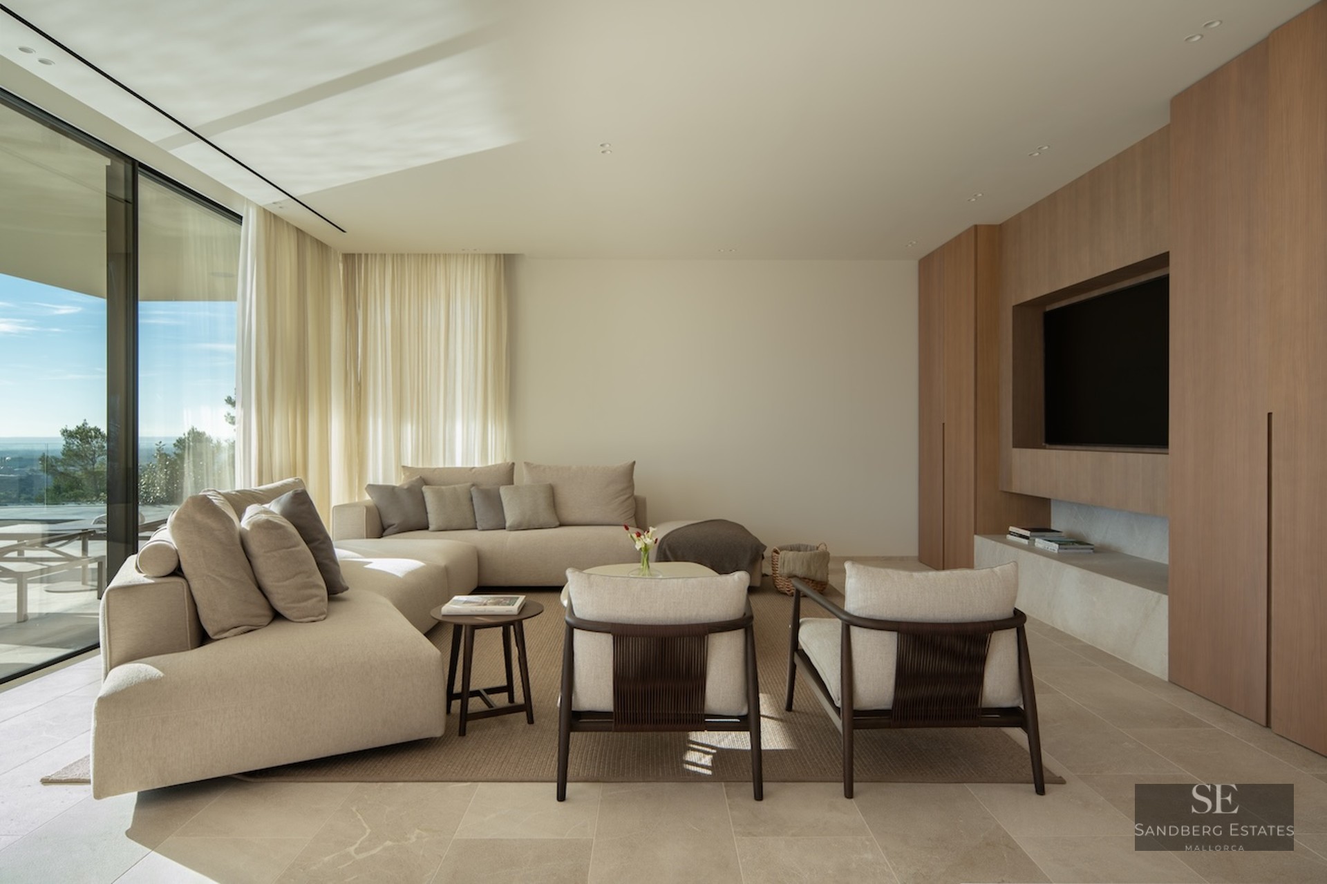 Modern living room with a beige sectional sofa, wood paneled TV wall, and floor-to-ceiling windows with a scenic view.