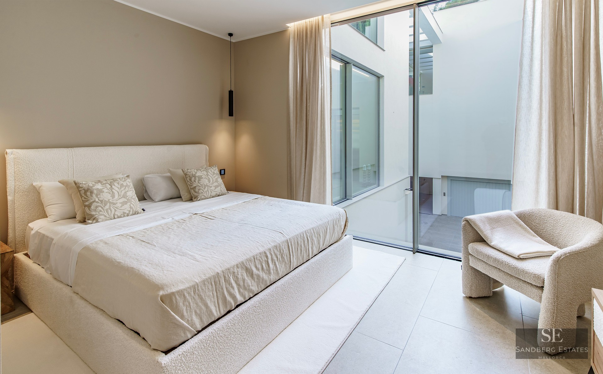 A bright, minimalist master bedroom with a cream boucle bed, matching armchair, and large windows facing an internal courtyard.