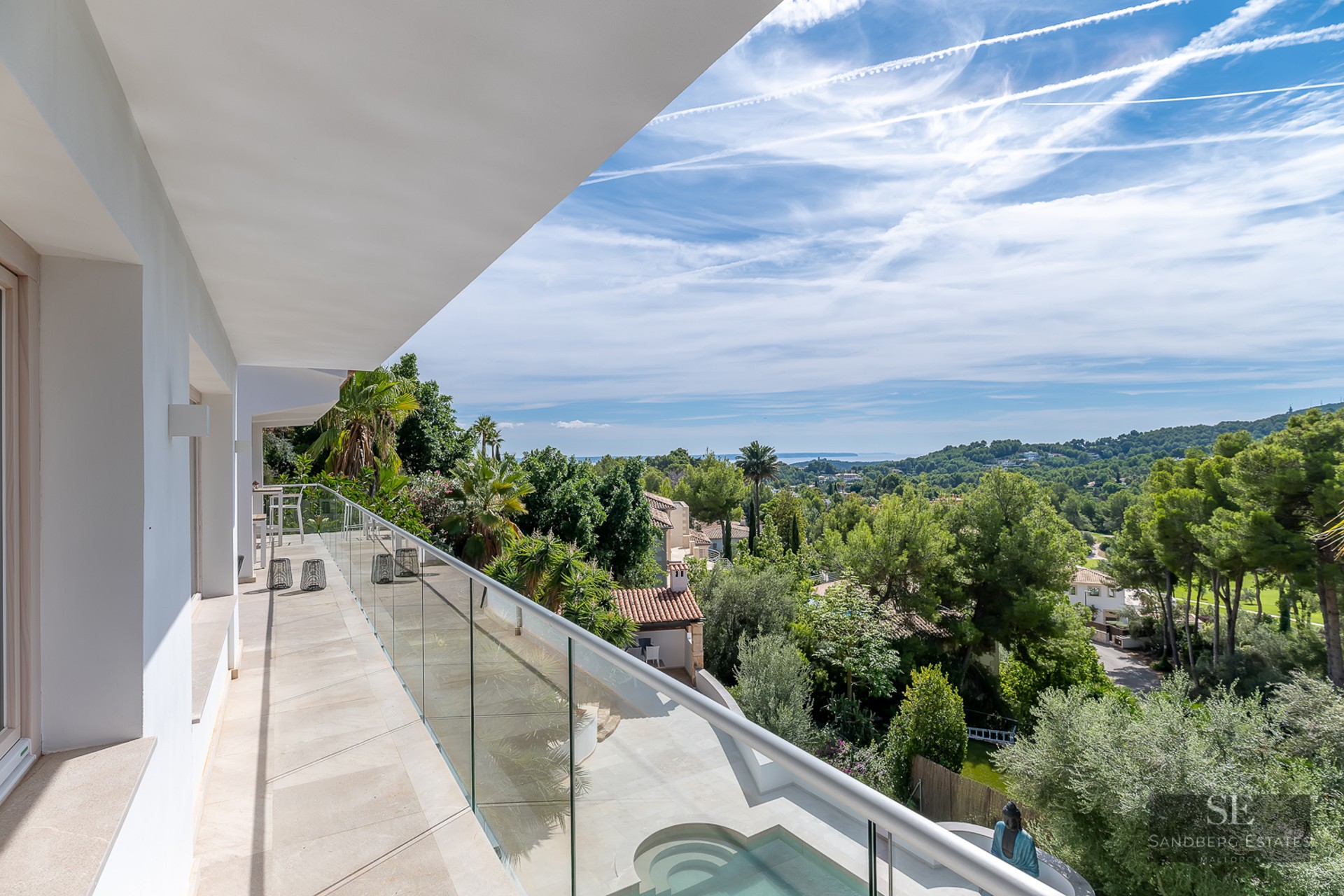 Balcony with glass railing overlooking lush green trees, a swimming pool, and the Mediterranean sea.