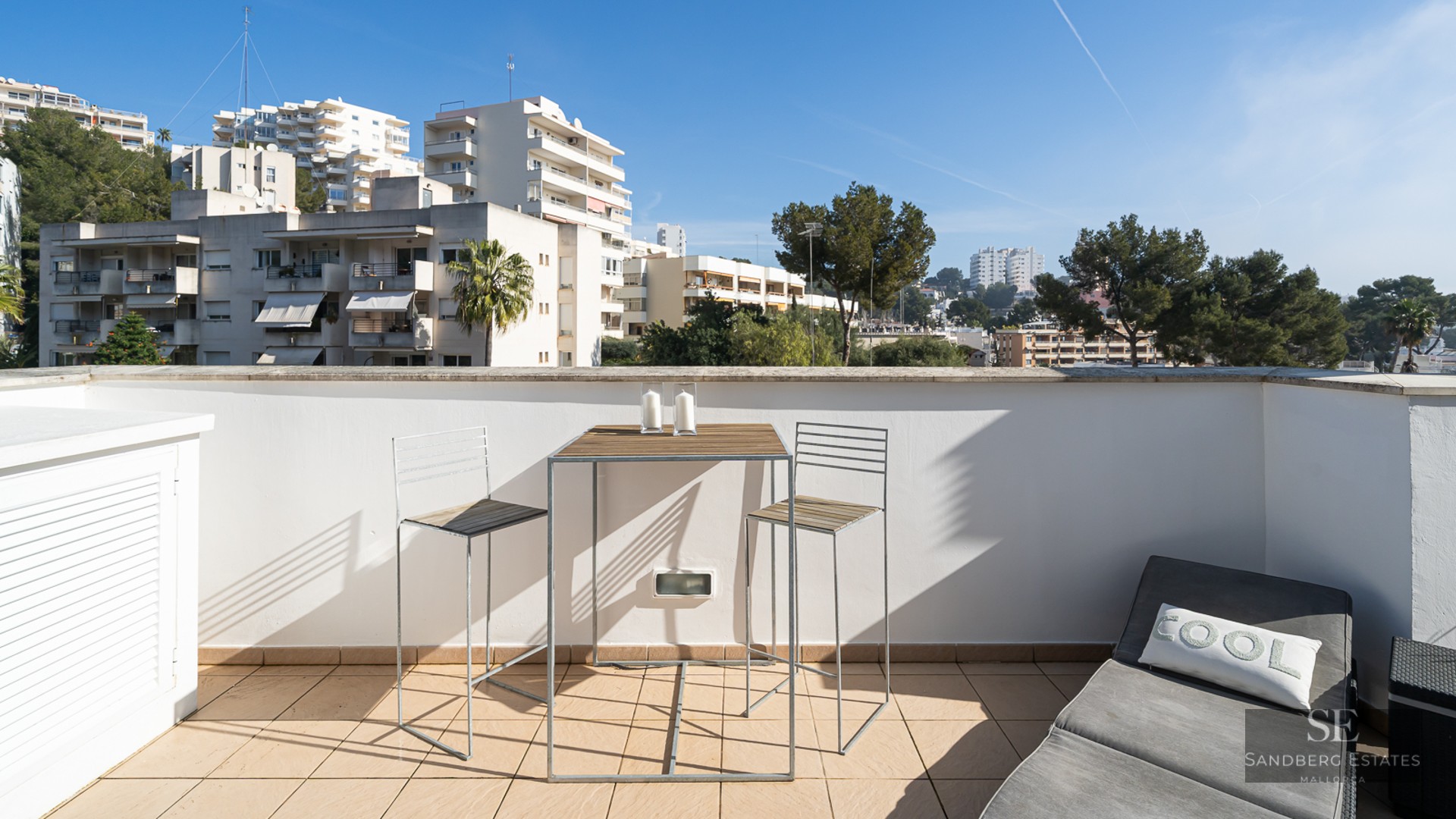 A sunlit terrace with a high-top wooden table, two bar chairs, and a grey sun lounger overlooking city buildings.