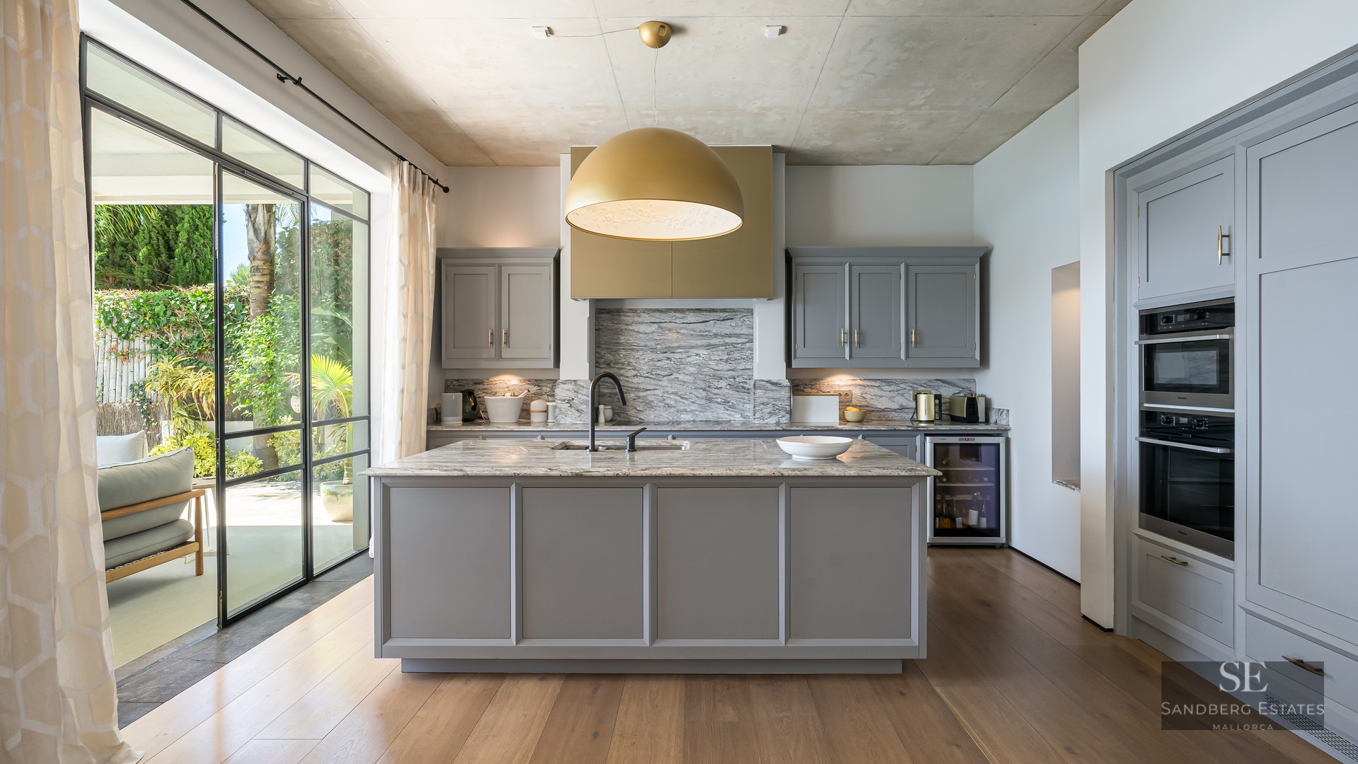 Modern kitchen with grey cabinets, marble island, gold pendant light, and floor-to-ceiling windows to a garden.