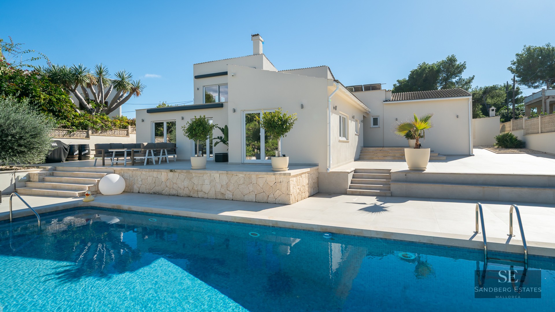 Piscine turquoise devant une villa blanche moderne avec terrasse en pierre sous un ciel bleu clair.