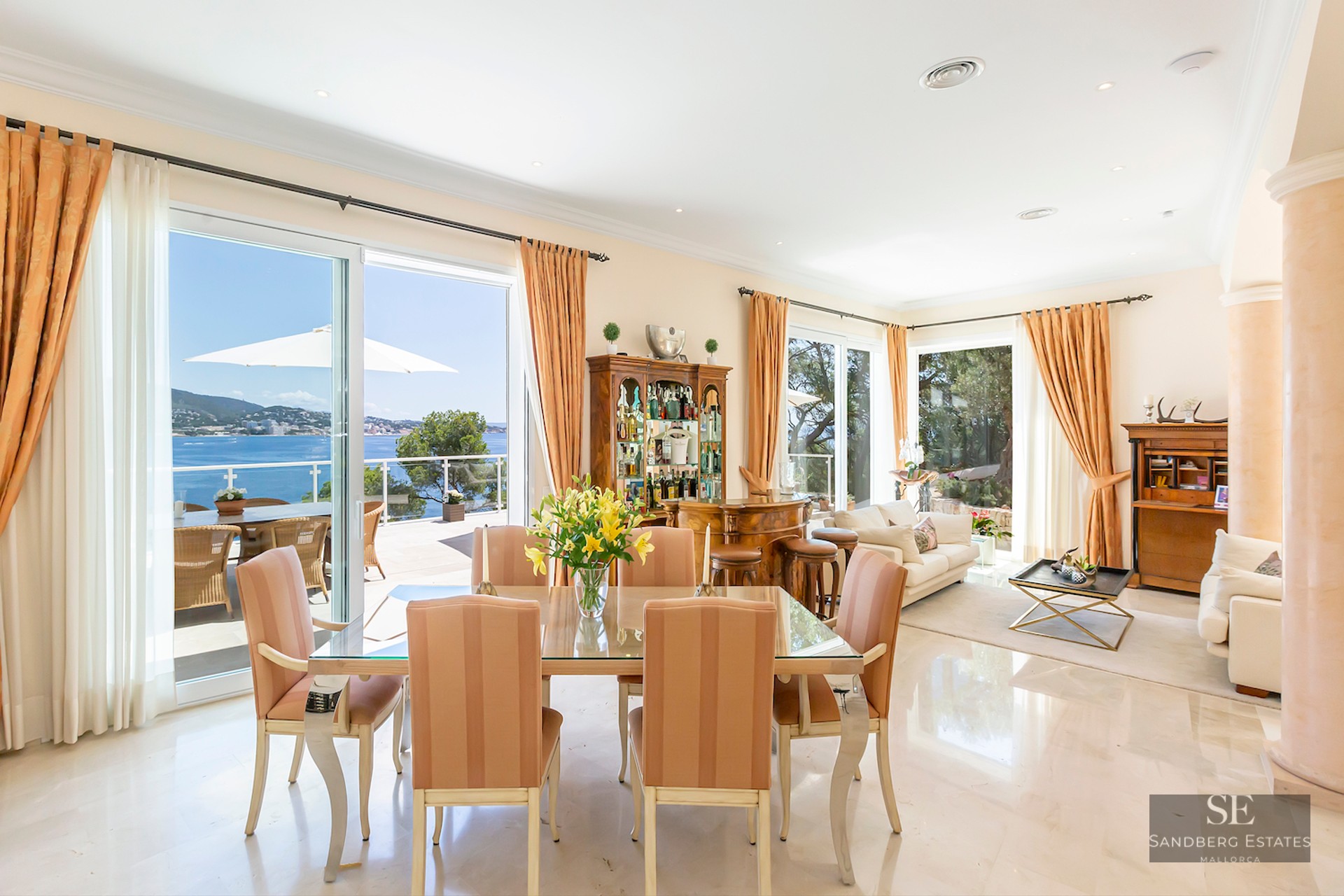 Bright open-plan dining room with a glass table, marble floors, and large windows overlooking the Mediterranean sea.