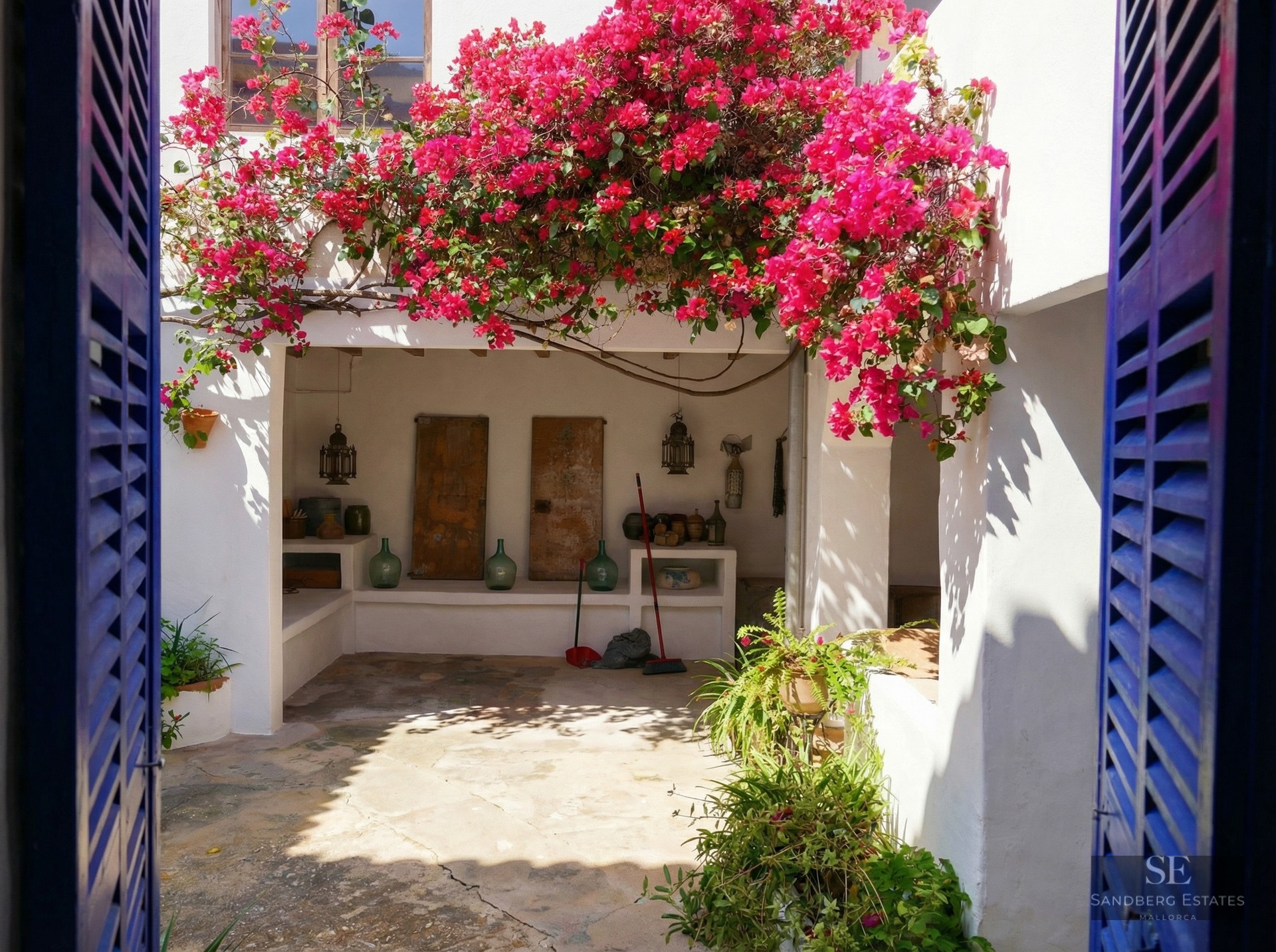 A sun-drenched white courtyard framed by blue shutters, featuring vibrant pink bougainvillea and rustic stone flooring.