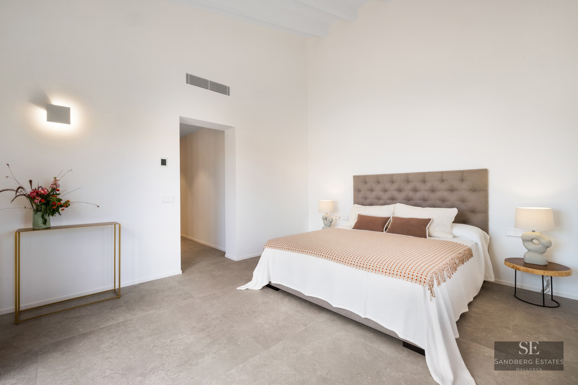 Minimalist bedroom with a tufted headboard, white bedding, designer lamps, and grey stone floors under high ceilings.