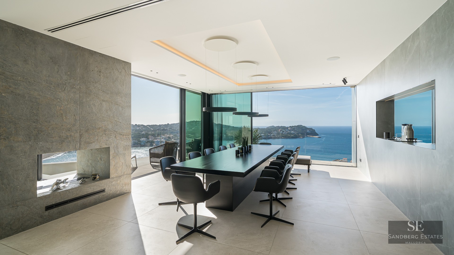 A modern dining room with a long black table, stone fireplace, and floor-to-ceiling windows overlooking the ocean.
