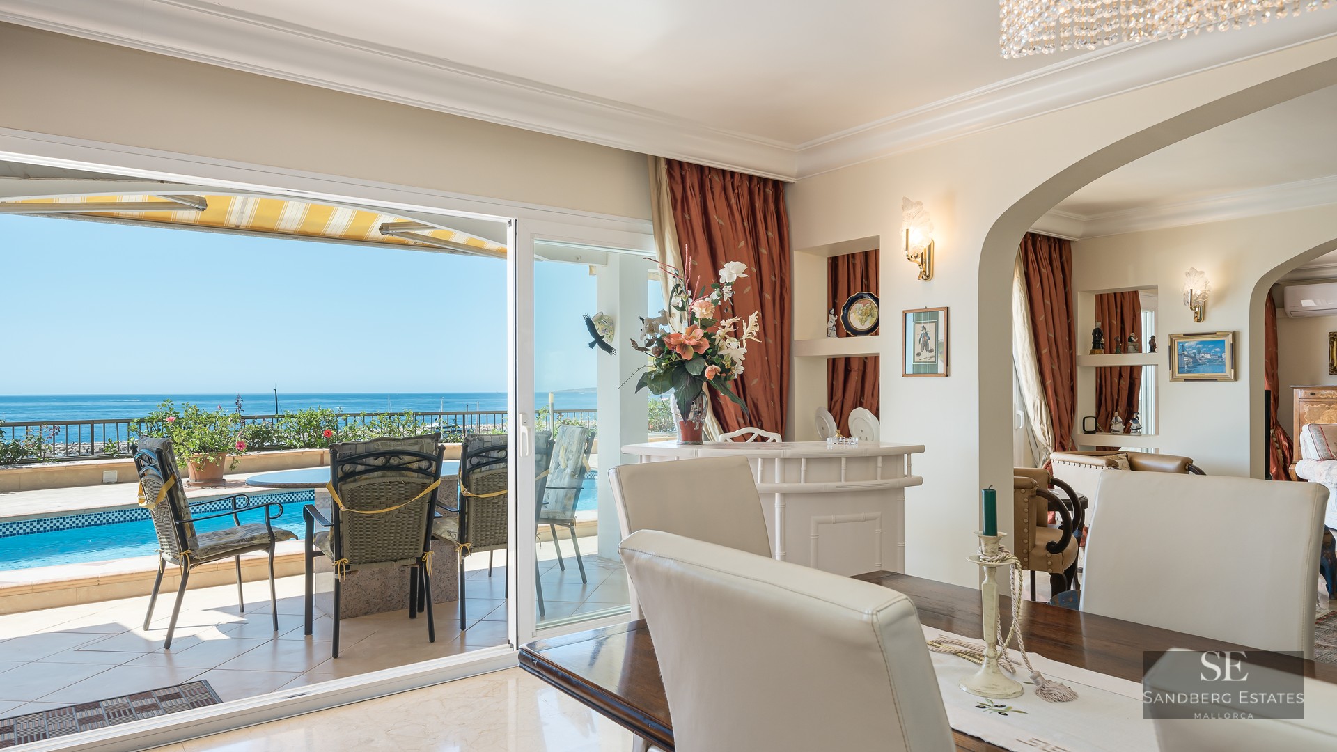 Interior dining area with a wooden table and white chairs looking out to a terrace with a pool and sea views.
