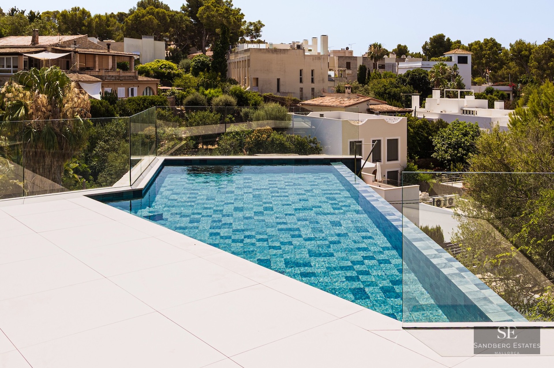 High-angle view of a triangular turquoise swimming pool with glass railings and a white tiled deck overlooking a town.
