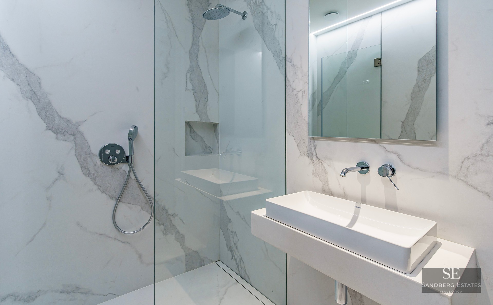 Modern bathroom with white marble-effect tiles, a glass walk-in shower, and a minimalist rectangular sink with chrome taps.