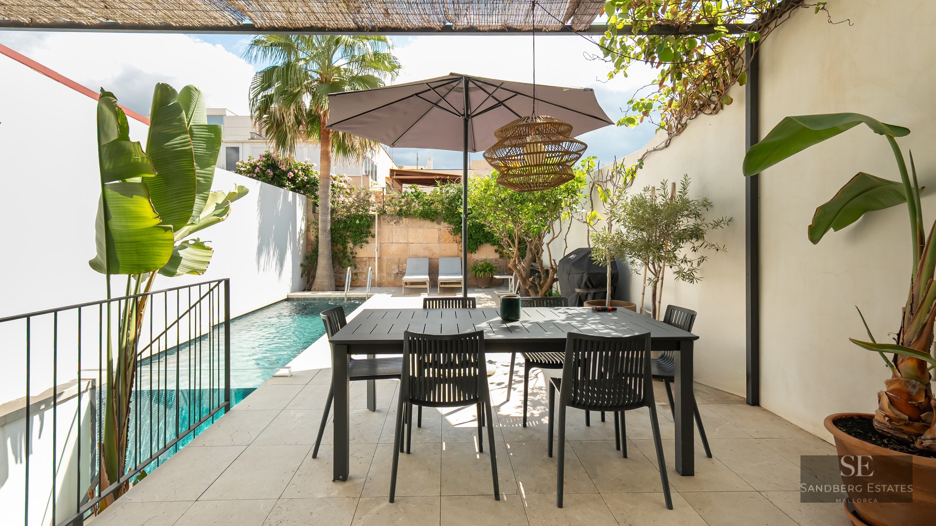 Modern outdoor terrace with a black dining table under an umbrella, next to a turquoise swimming pool and palm trees.