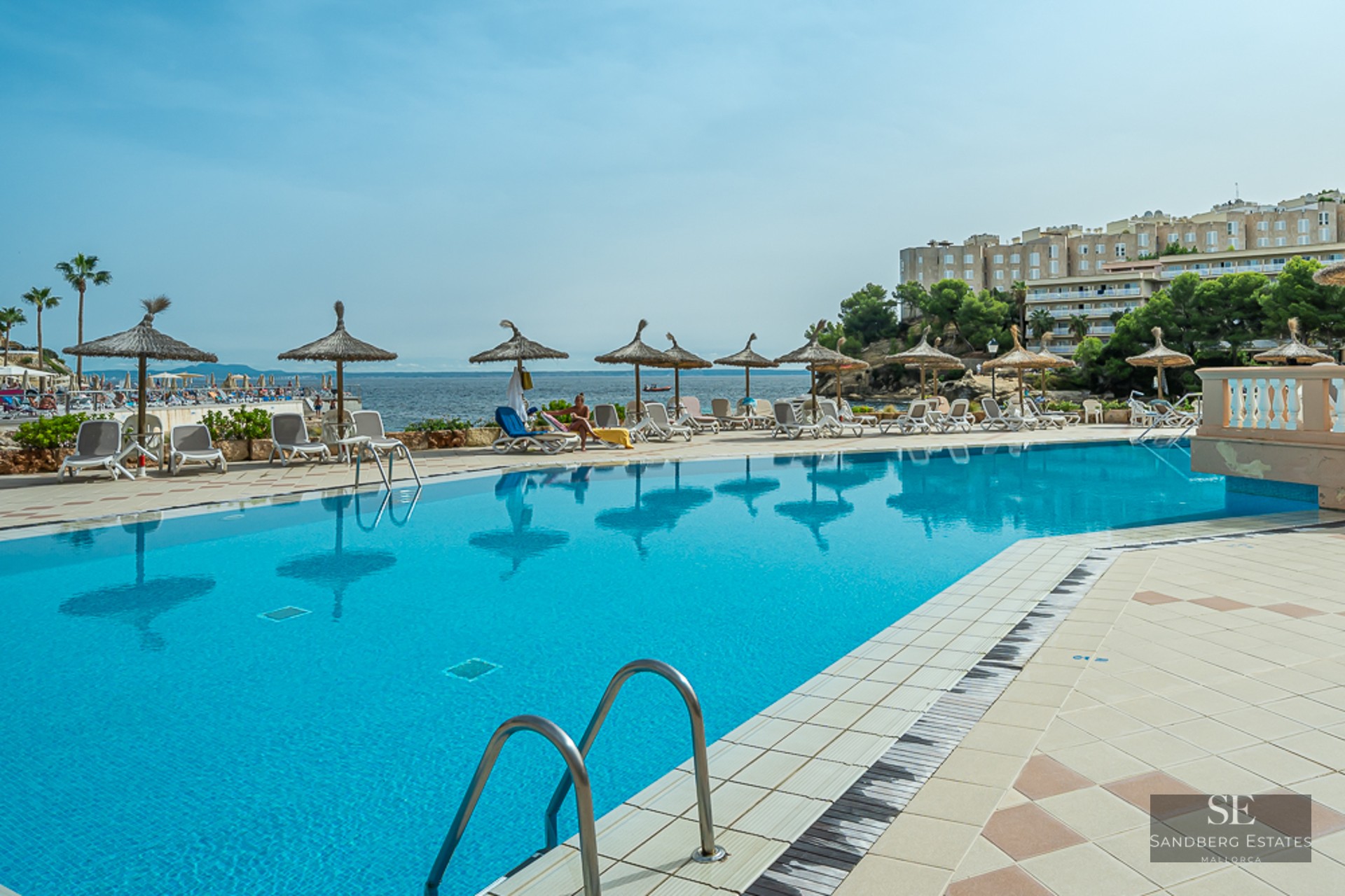 Large blue swimming pool surrounded by sunbeds and straw umbrellas, overlooking the sea and coastal buildings.