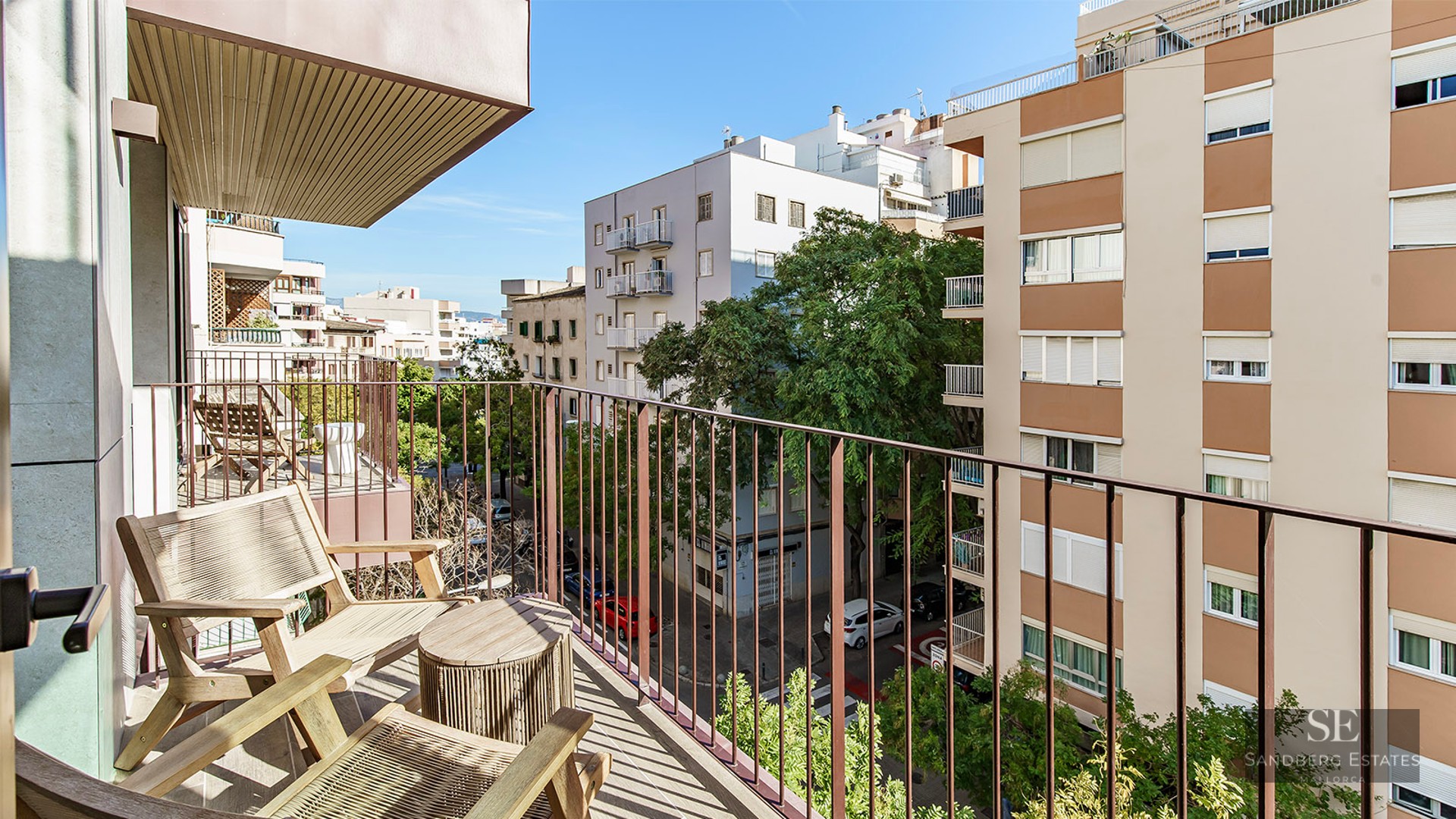 A sunny balcony with wooden chairs and a table overlooking a tree-lined city street and apartment buildings.