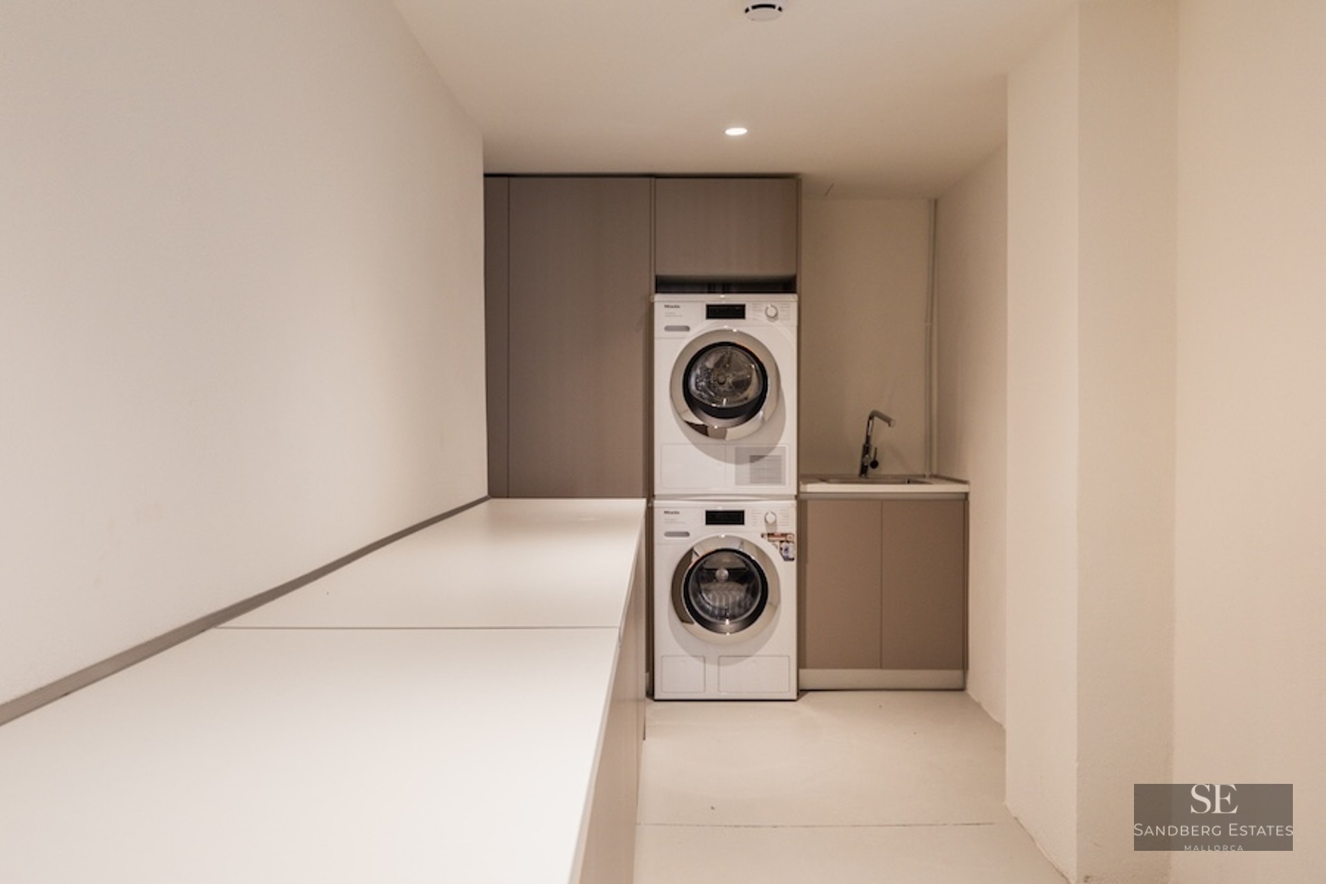 Stacked Miele washer and dryer in a minimalist laundry room with beige cabinetry and a long white countertop.