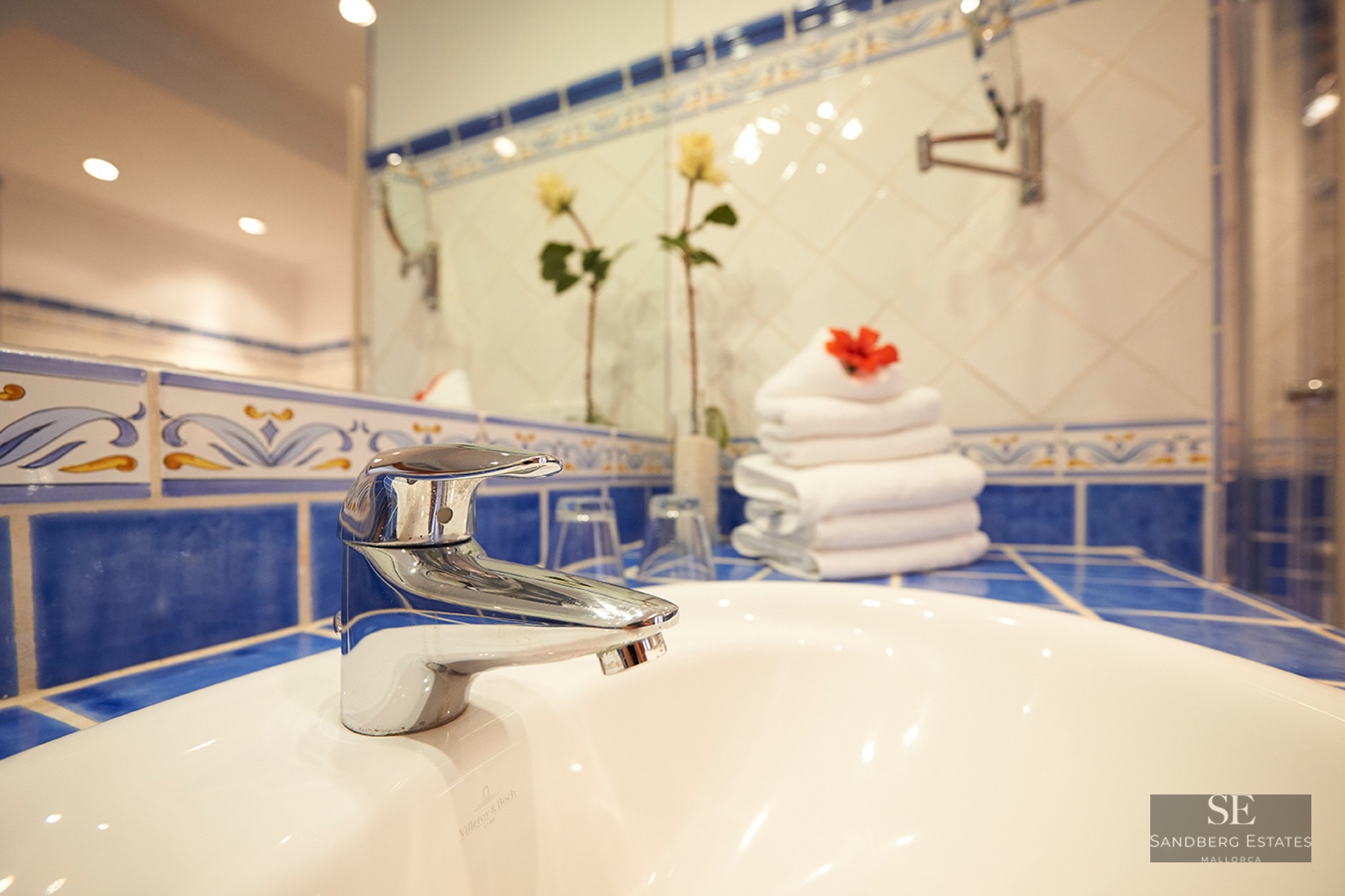 Close-up of a white porcelain sink and chrome faucet with blue patterned Mediterranean tiles and white towels.