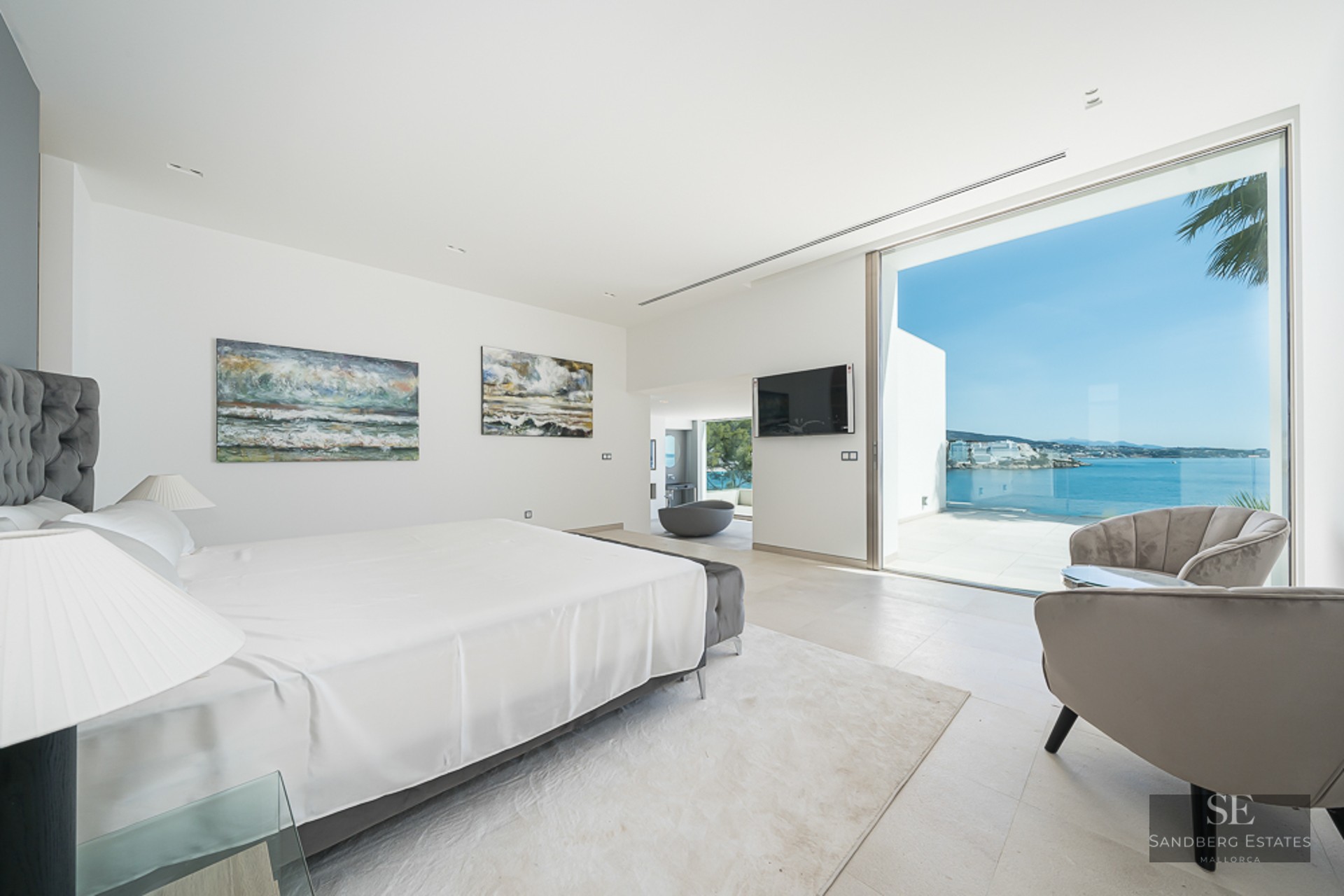 Bright master bedroom with white bedding, grey velvet chairs, and large glass doors overlooking the Mediterranean Sea.