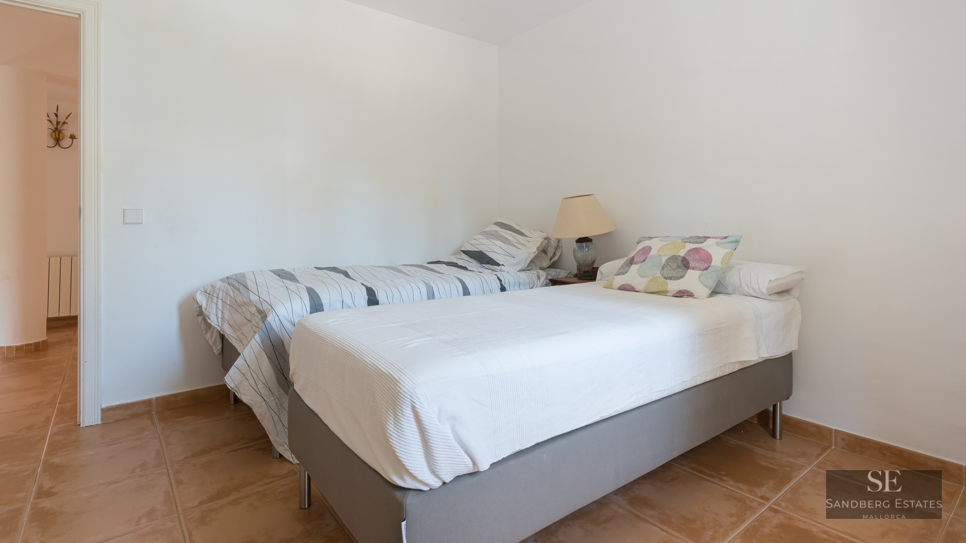 Simple bedroom featuring two single beds, terracotta tile flooring, and clean white walls.