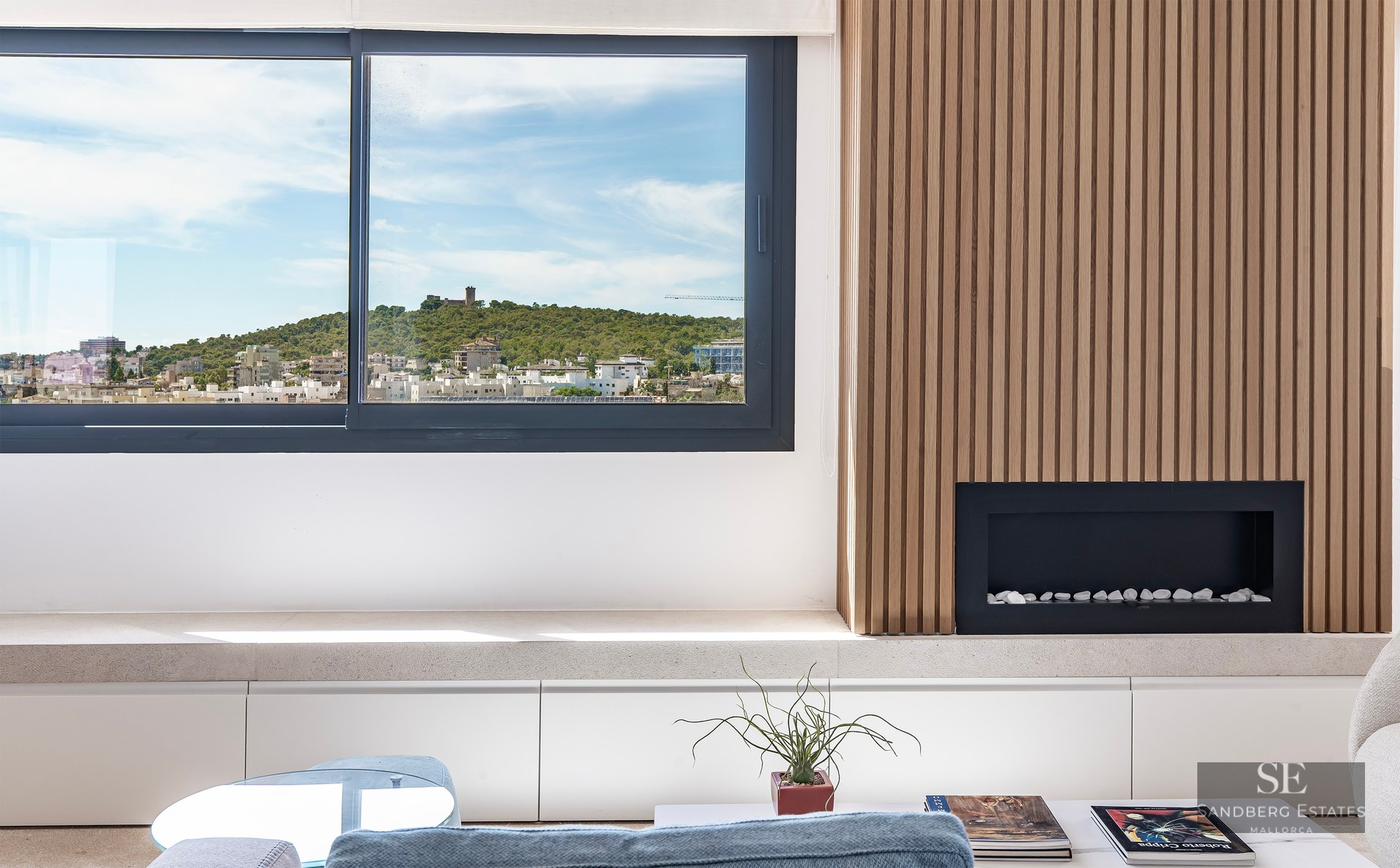 Modern living room with slatted wood wall, built-in fireplace, and a large window overlooking a hilltop castle.