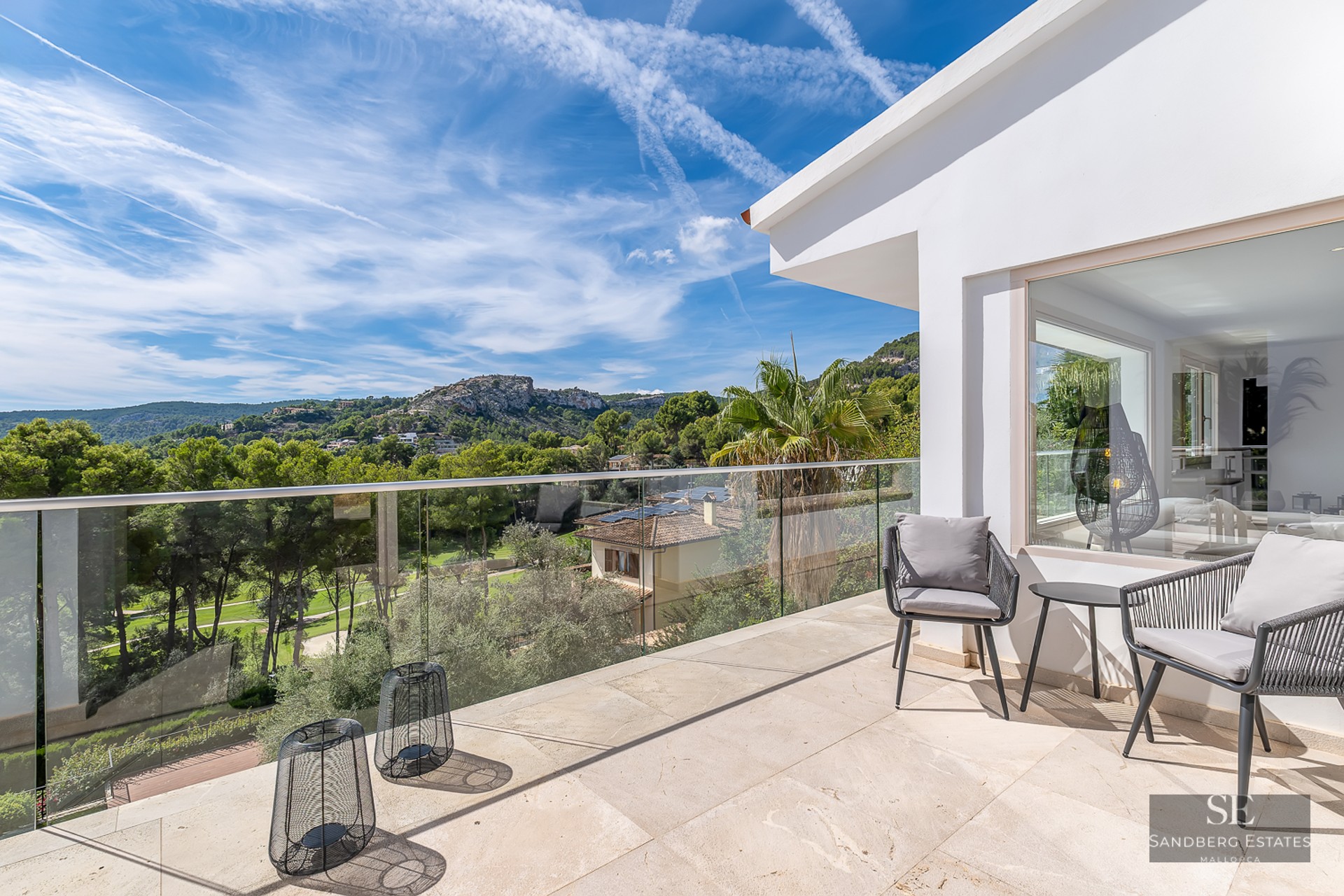 Spacious sun-drenched terrace with glass railings, modern grey chairs, and a breathtaking view of green hills.