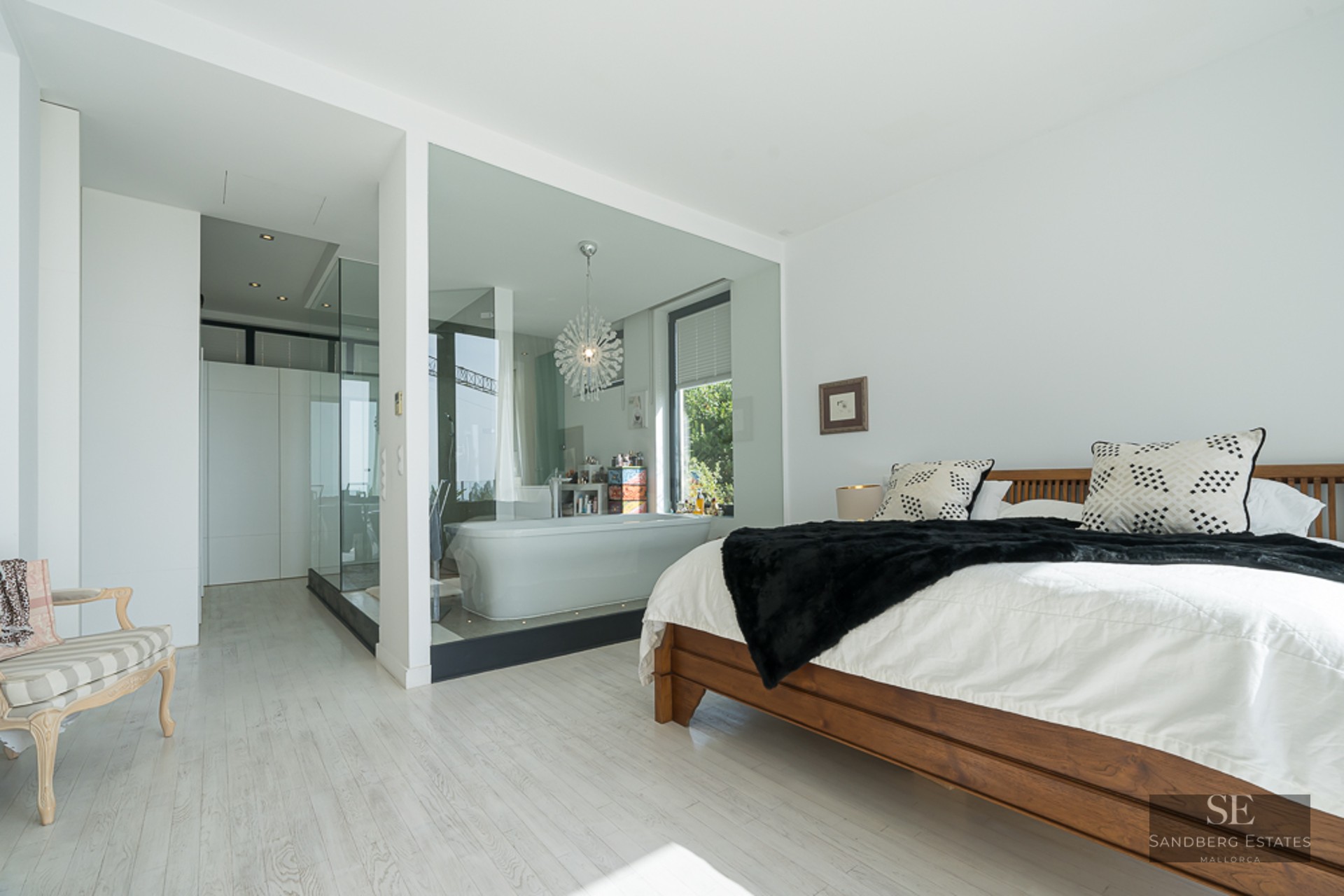 Modern master bedroom featuring a wooden bed and a glass-walled bathroom with a freestanding tub and designer chandelier.