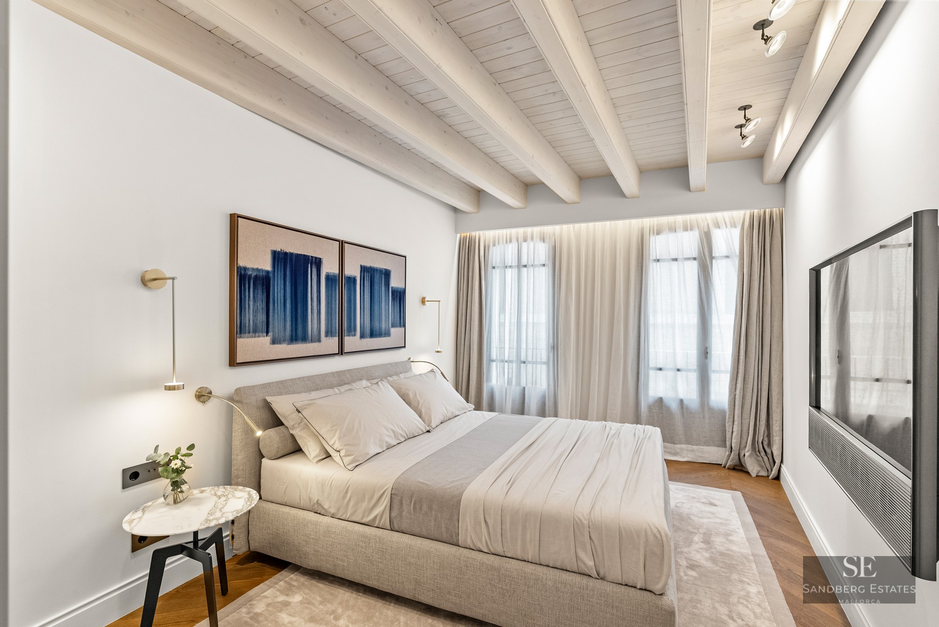 Modern bedroom with wooden ceiling beams, grey upholstered bed, marble side table, and floor-to-ceiling curtains.