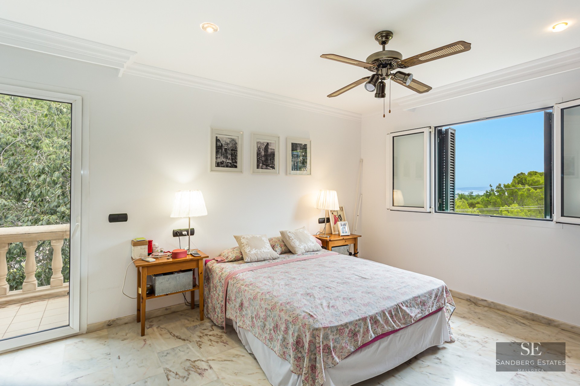 White bedroom with marble floor, double bed, ceiling fan, and window showing a scenic sea and forest view.