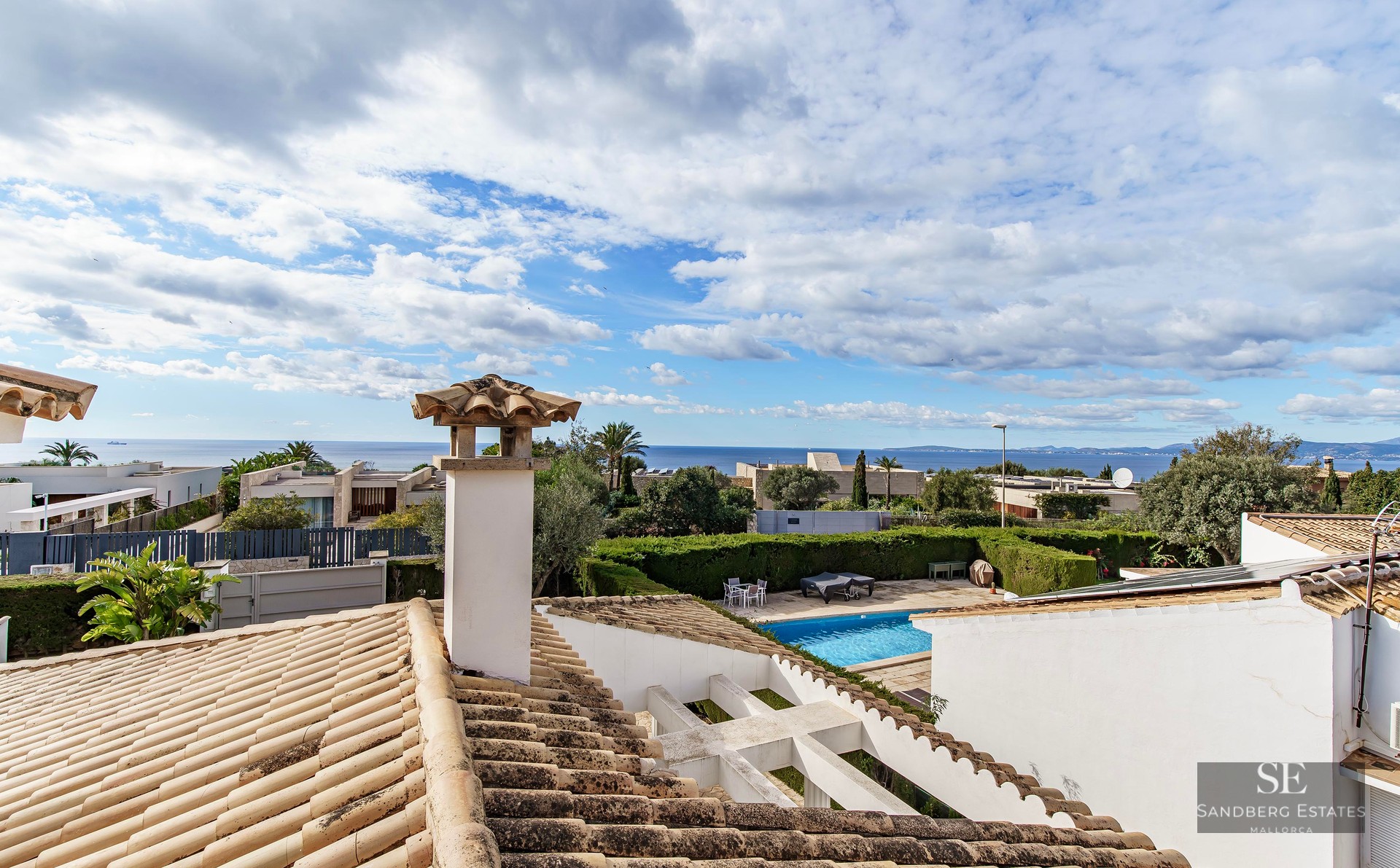 View from a tiled rooftop looking over a garden pool toward the Mediterranean Sea under a cloudy sky.