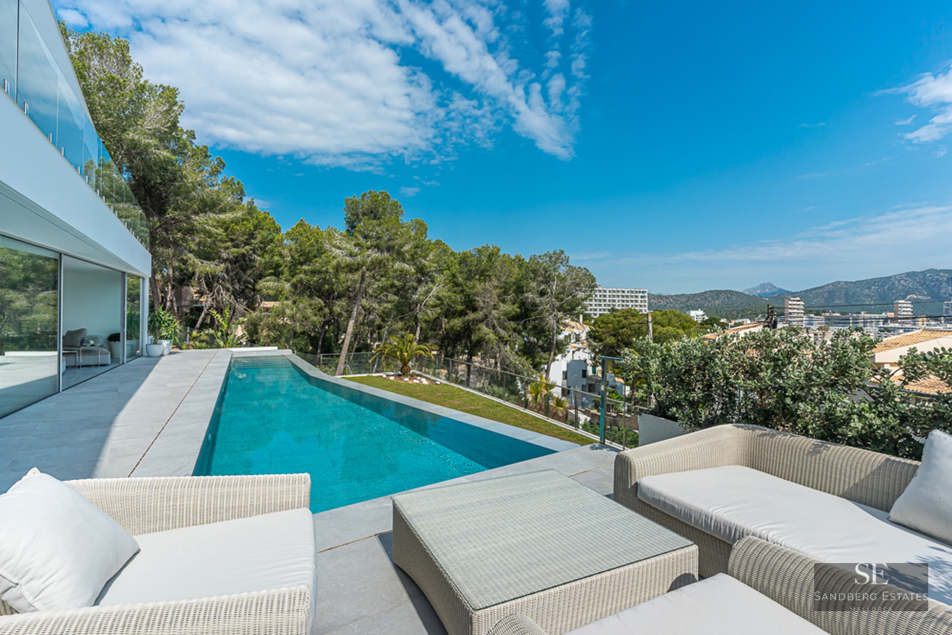 Modern terrace with a long blue swimming pool, white wicker lounge furniture, glass railings, and forest views.
