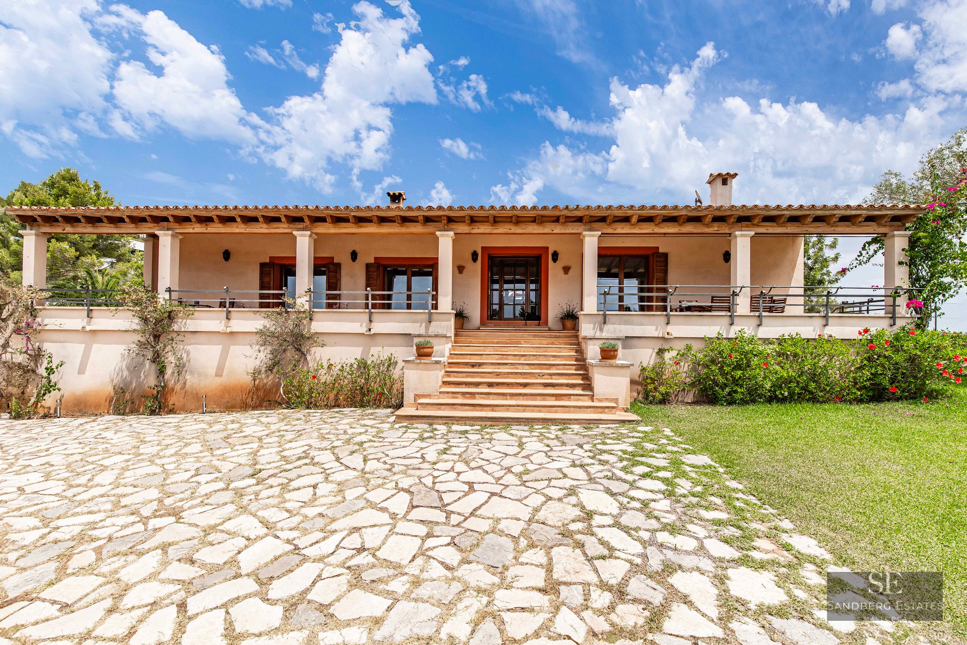 Front view of a one-story Mediterranean villa with a stone driveway, steps, and a covered terrace under a blue sky.