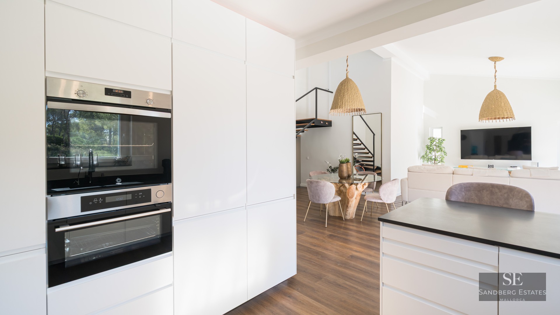 Cuisine blanche minimaliste avec fours encastrés donnant sur une salle à manger avec table en verre et chaises roses.