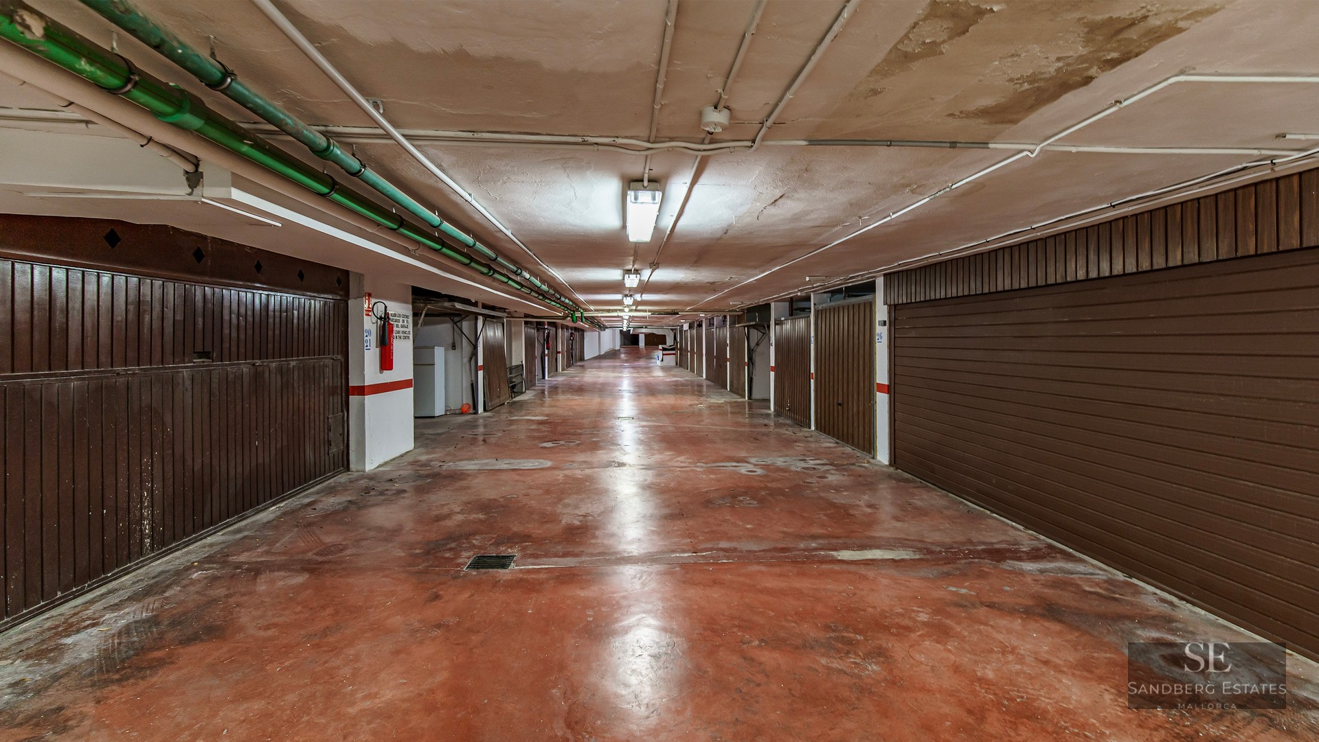 Underground garage hallway with red concrete floor and individual brown garage doors on both sides.