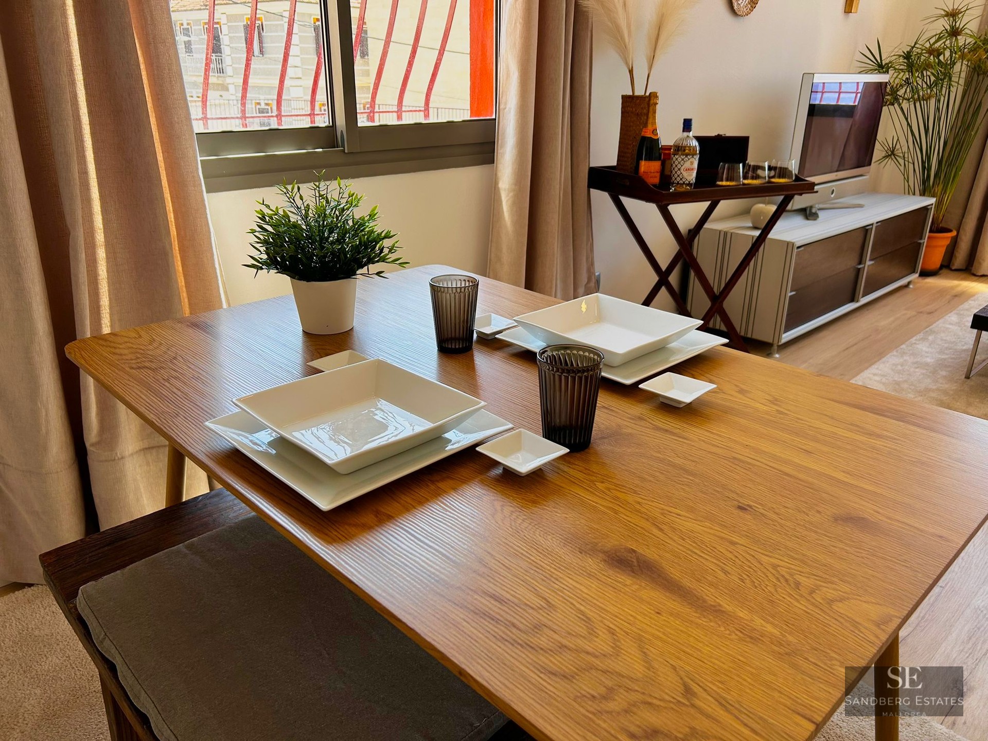 Wooden dining table set with white square plates and glasses in a bright living room.
