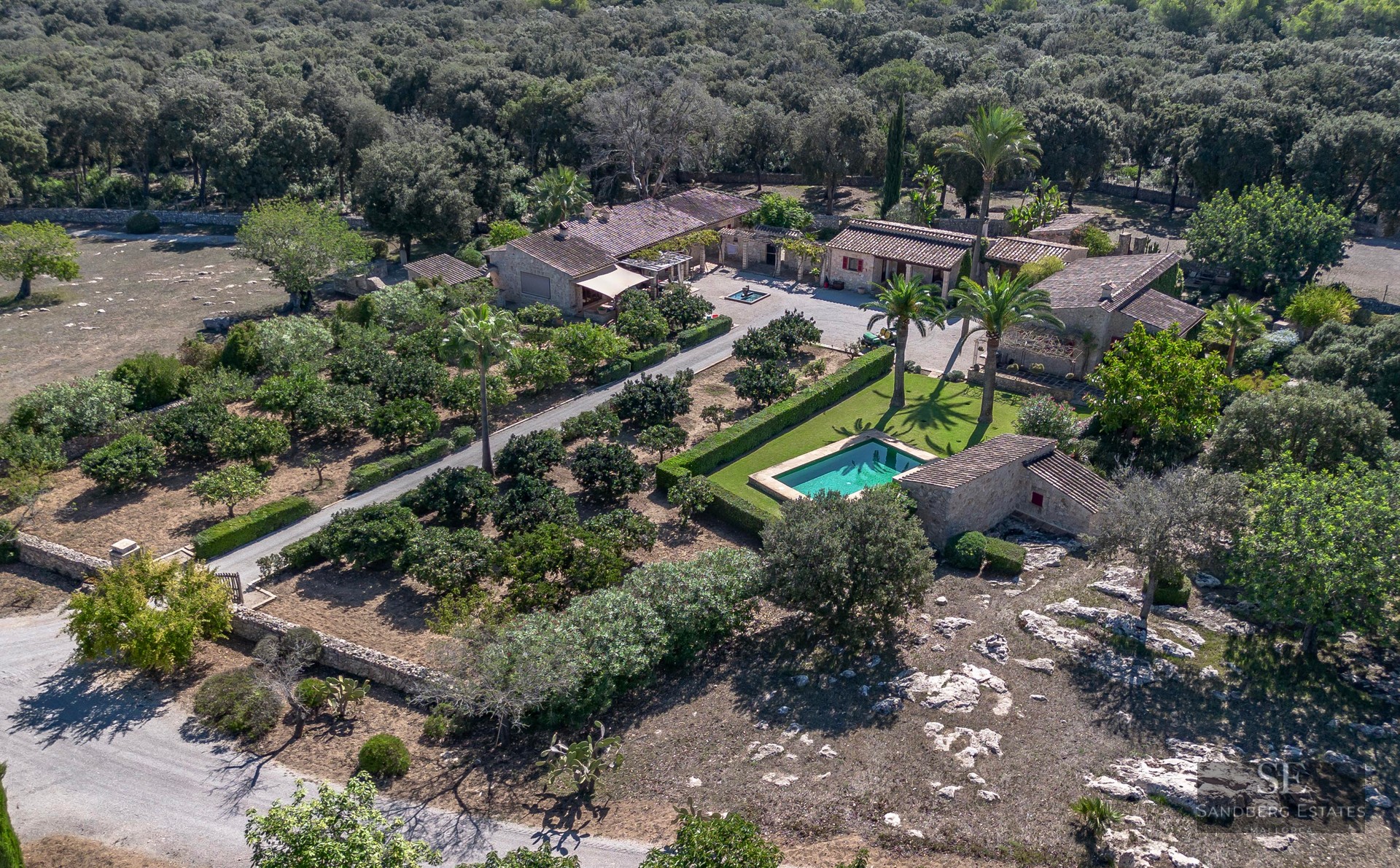 Bird's eye view of a stone villa estate with a swimming pool, palm trees, and lush gardens surrounded by forest.