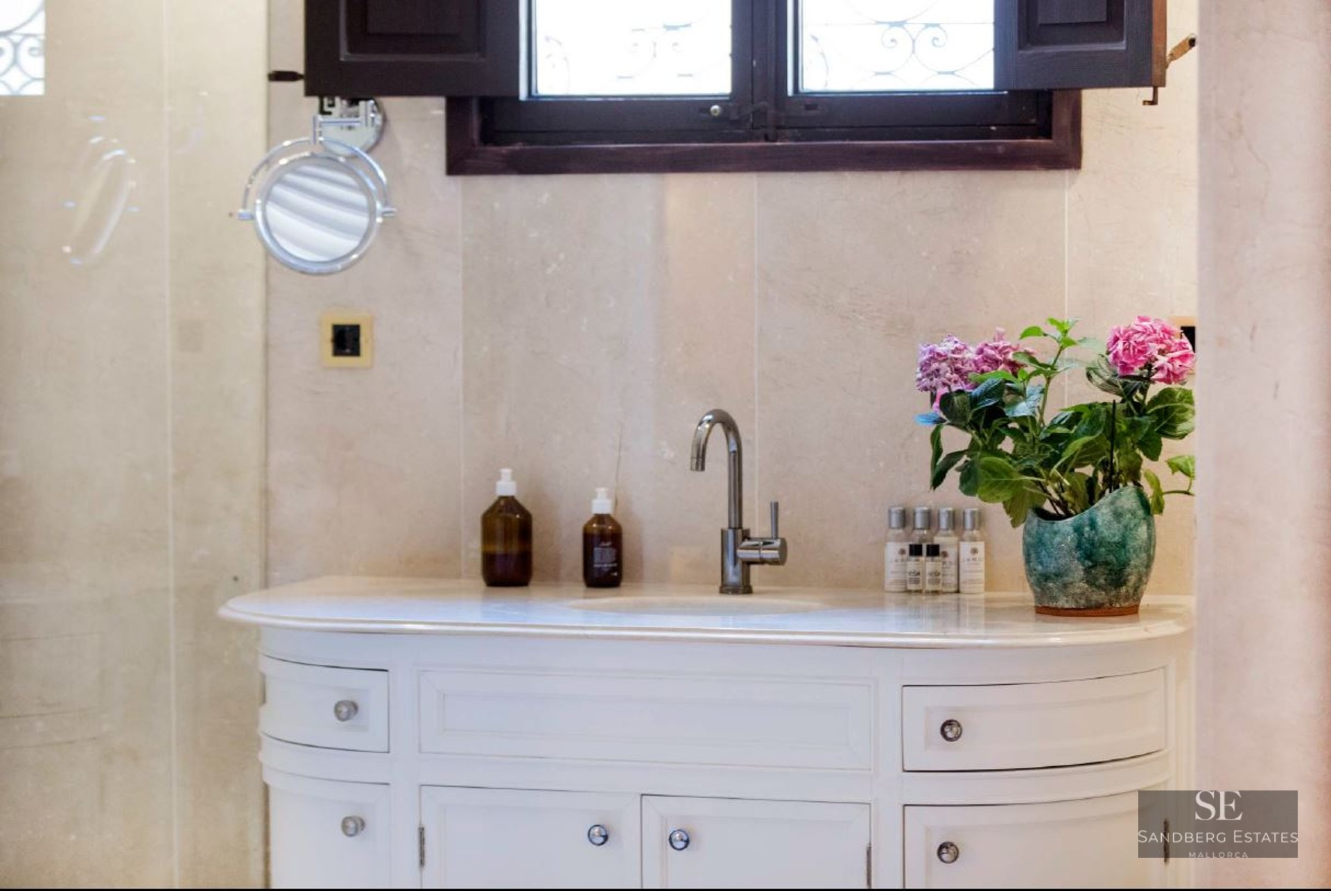 White vanity with chrome faucet, beige marble walls, magnifying mirror, and pink flowers in a pot.