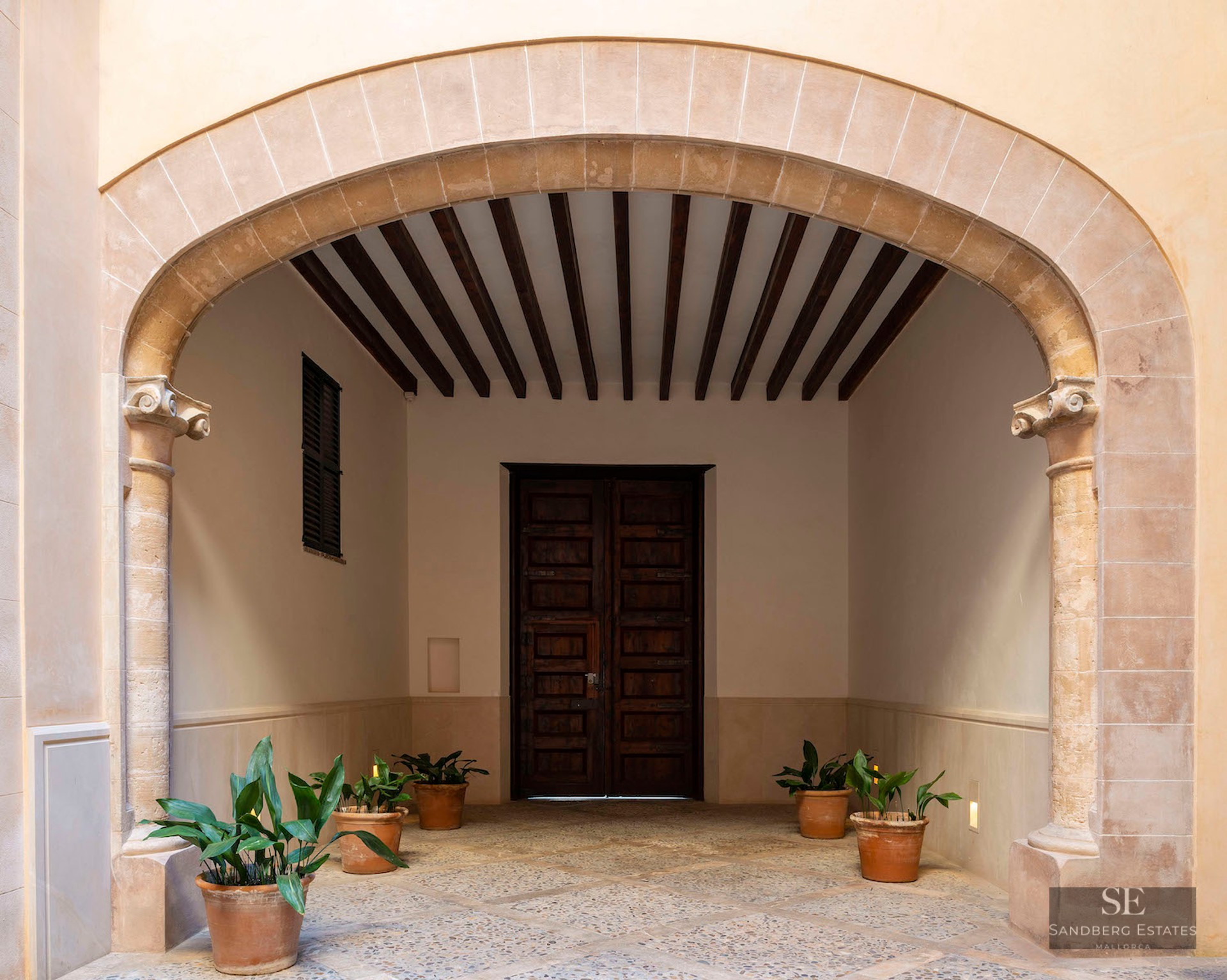 A grand stone archway leads to a traditional wooden door with exposed ceiling beams and potted plants.
