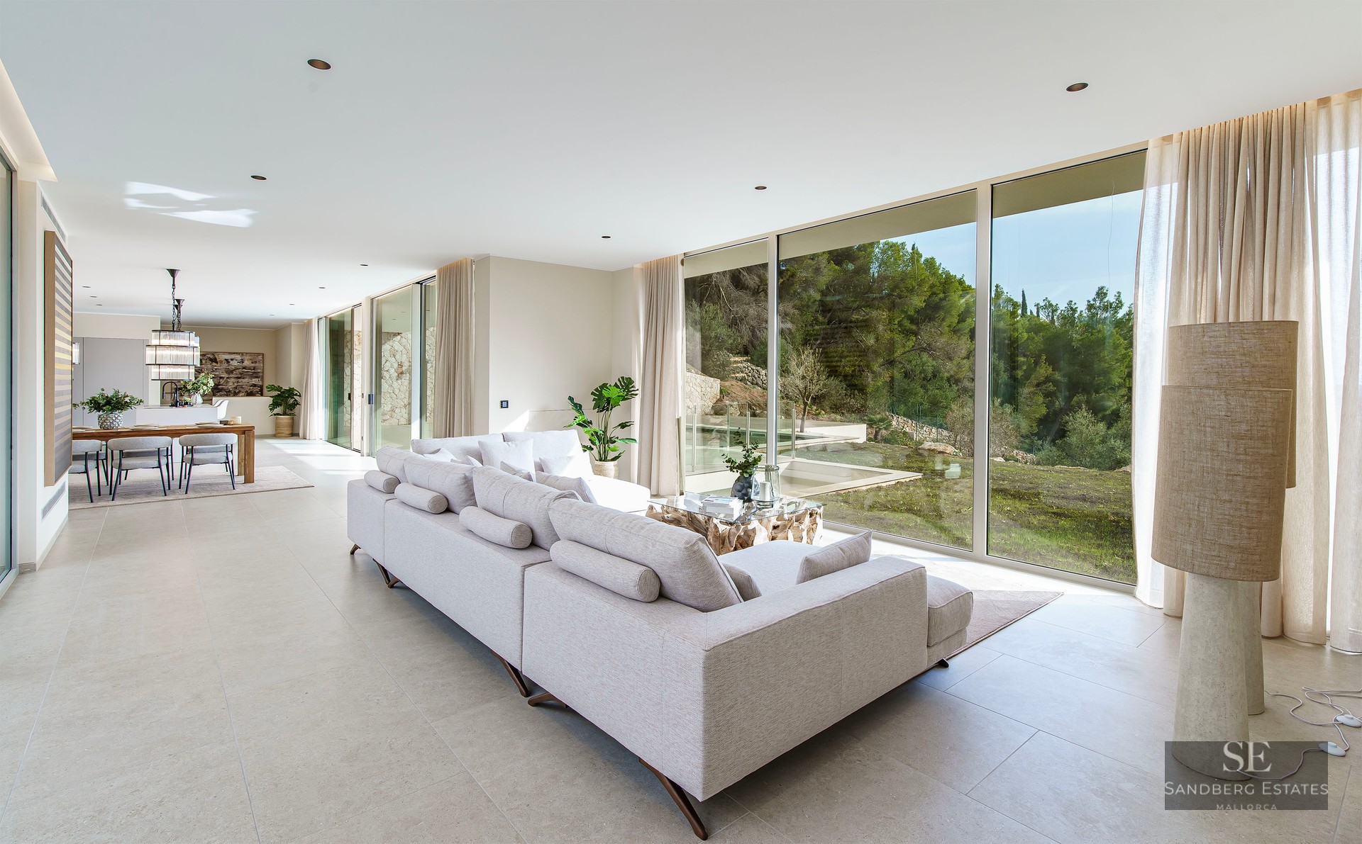 Spacious modern living room with a large grey sofa and floor-to-ceiling windows overlooking a lush green forest.