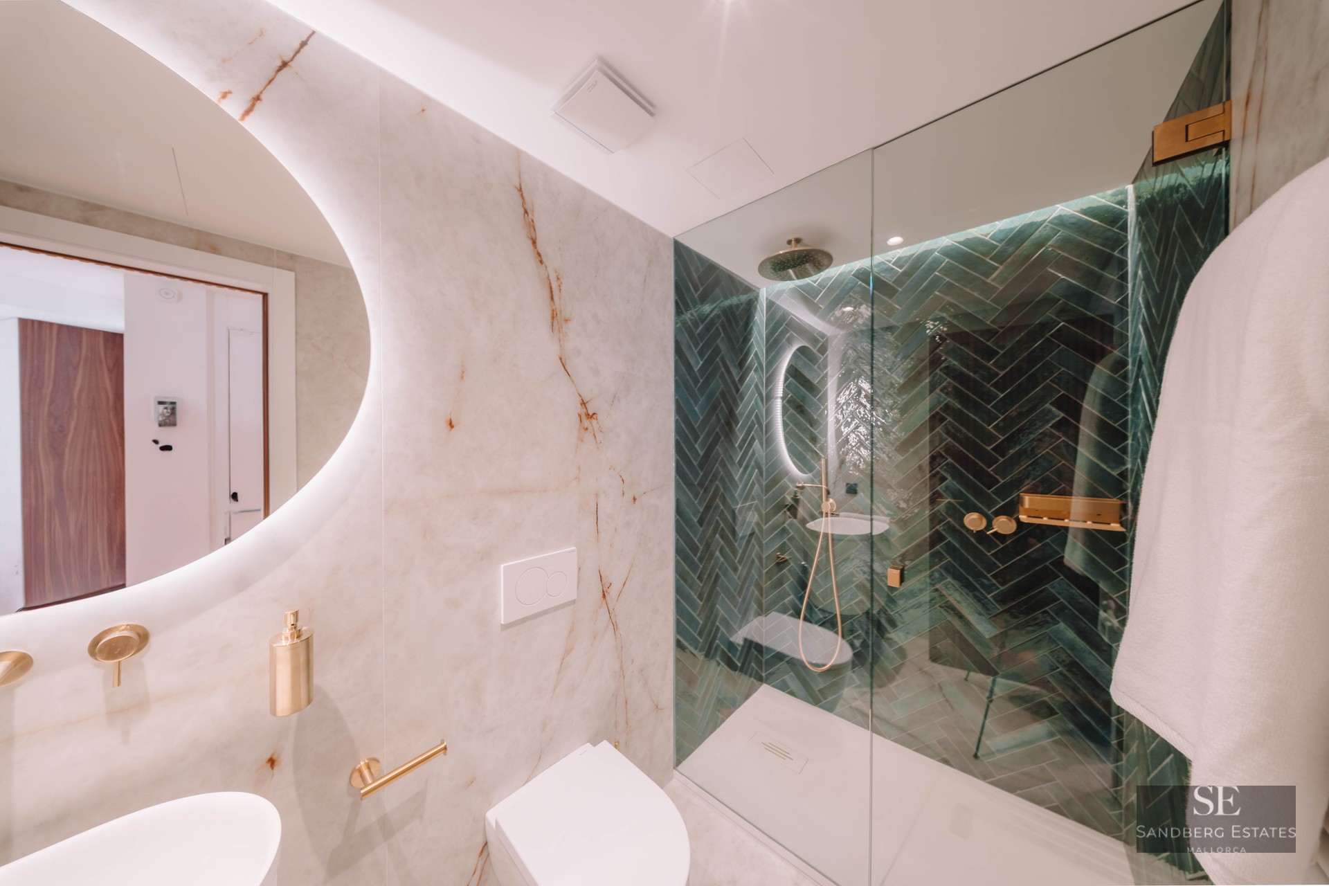 Modern bathroom with white marble walls, a round backlit mirror, and a walk-in shower with green herringbone tiles.