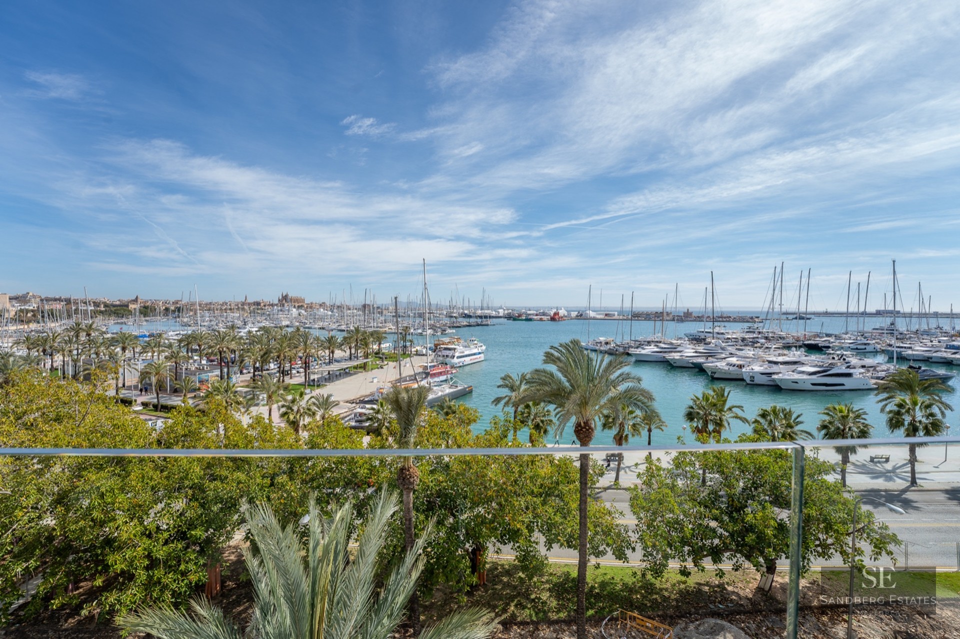 Elevated view through a glass railing overlooking a yacht-filled marina with palm trees and a cathedral in the distance.