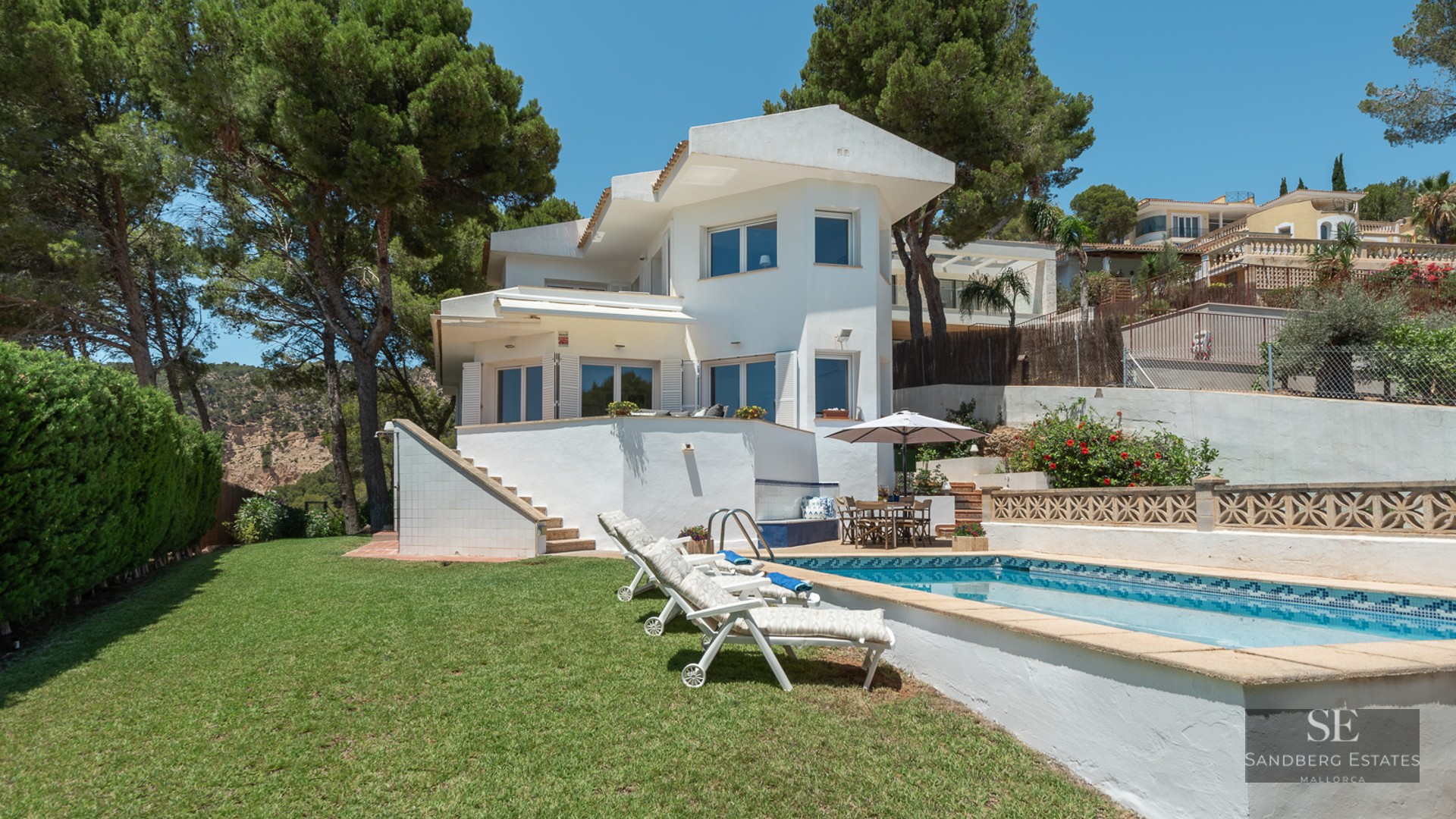 White two-story villa with a swimming pool, loungers, and green lawn surrounded by tall pine trees.