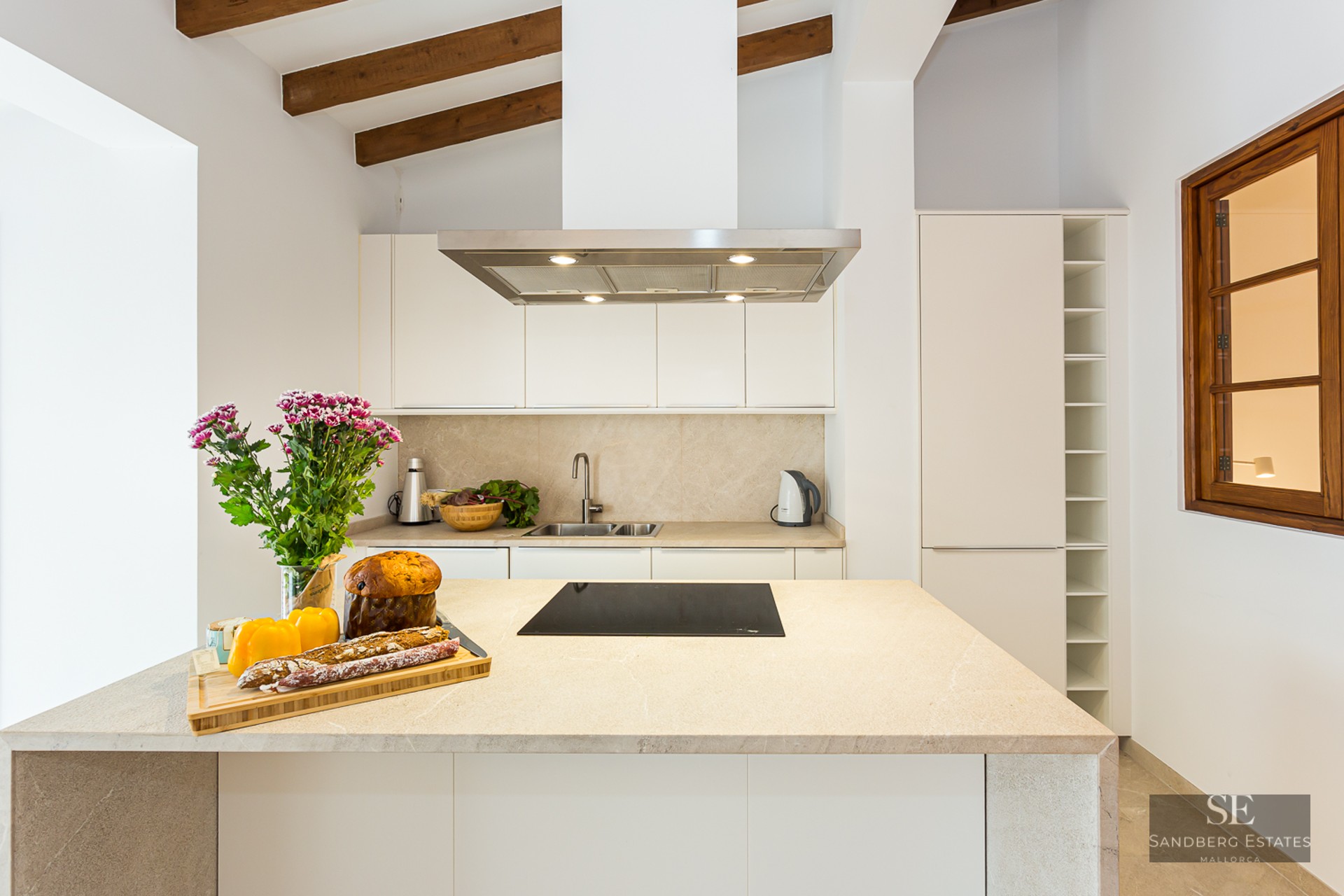 Bright kitchen featuring white cabinetry, a large stone island, rustic wooden ceiling beams, and integrated appliances.