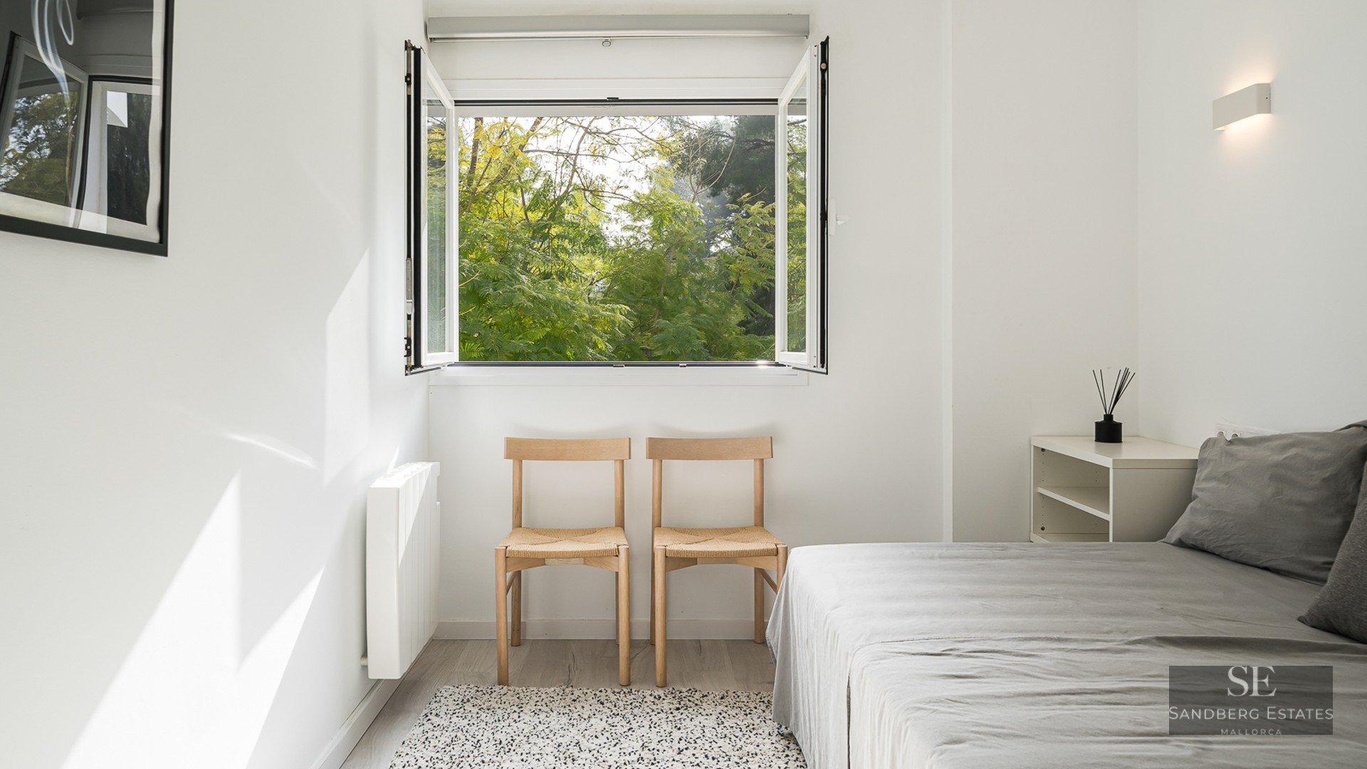 Bright minimalist bedroom with white walls, wooden chairs, grey bed, and an open window viewing lush green trees.