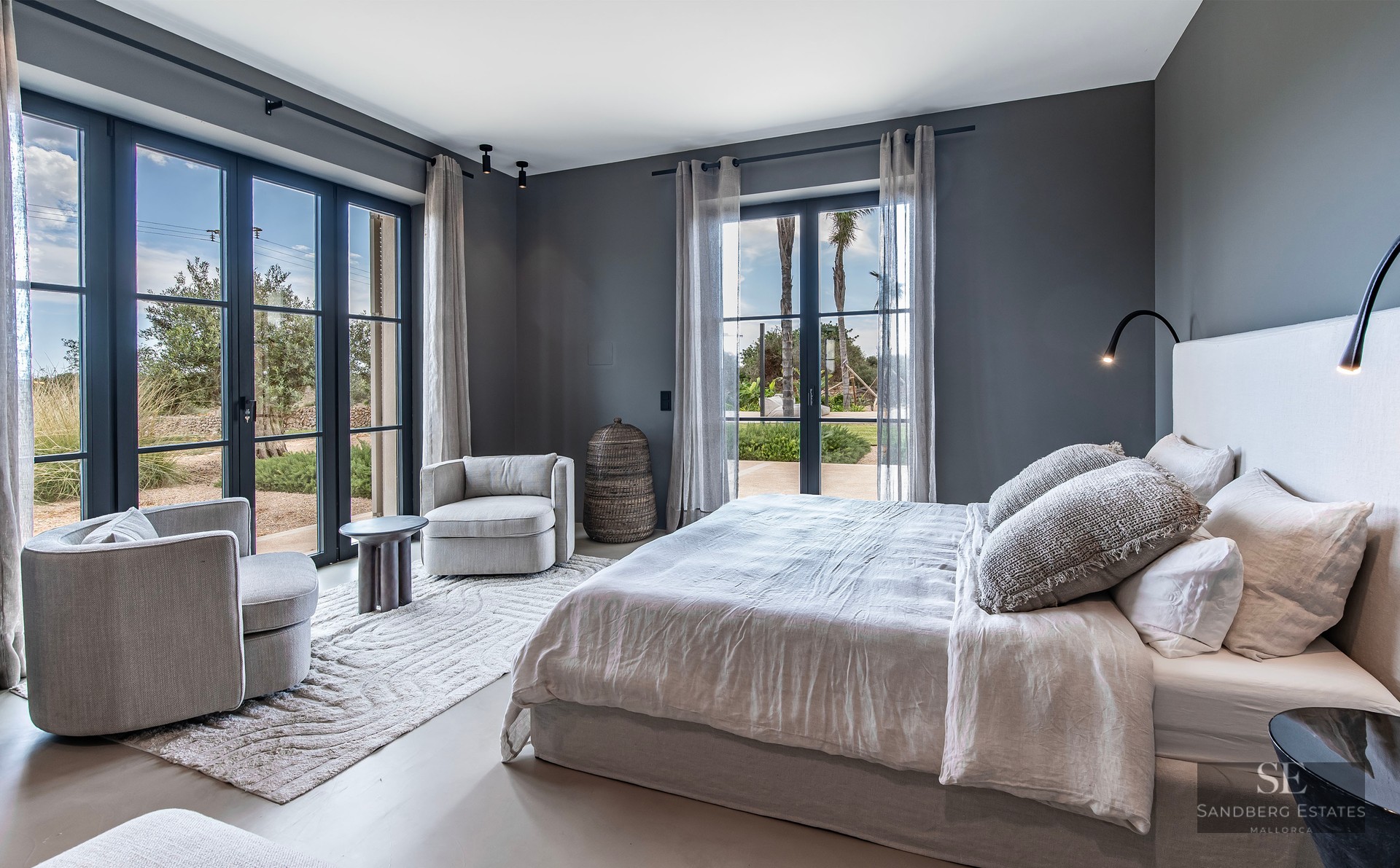Elegant bedroom with grey walls, a large bed, armchairs, and floor-to-ceiling windows opening to a garden.
