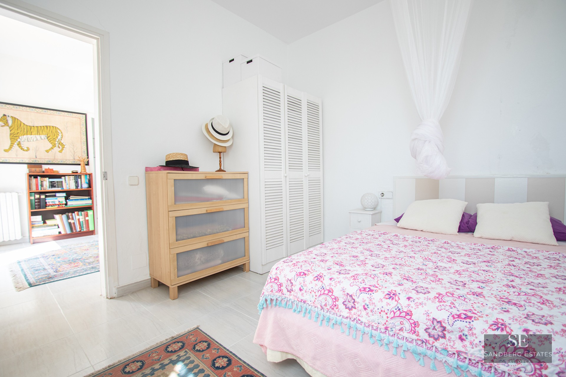 Bedroom with pink patterned bedspread, white canopy, light wood dresser, and white louvered wardrobe on tiled floor.