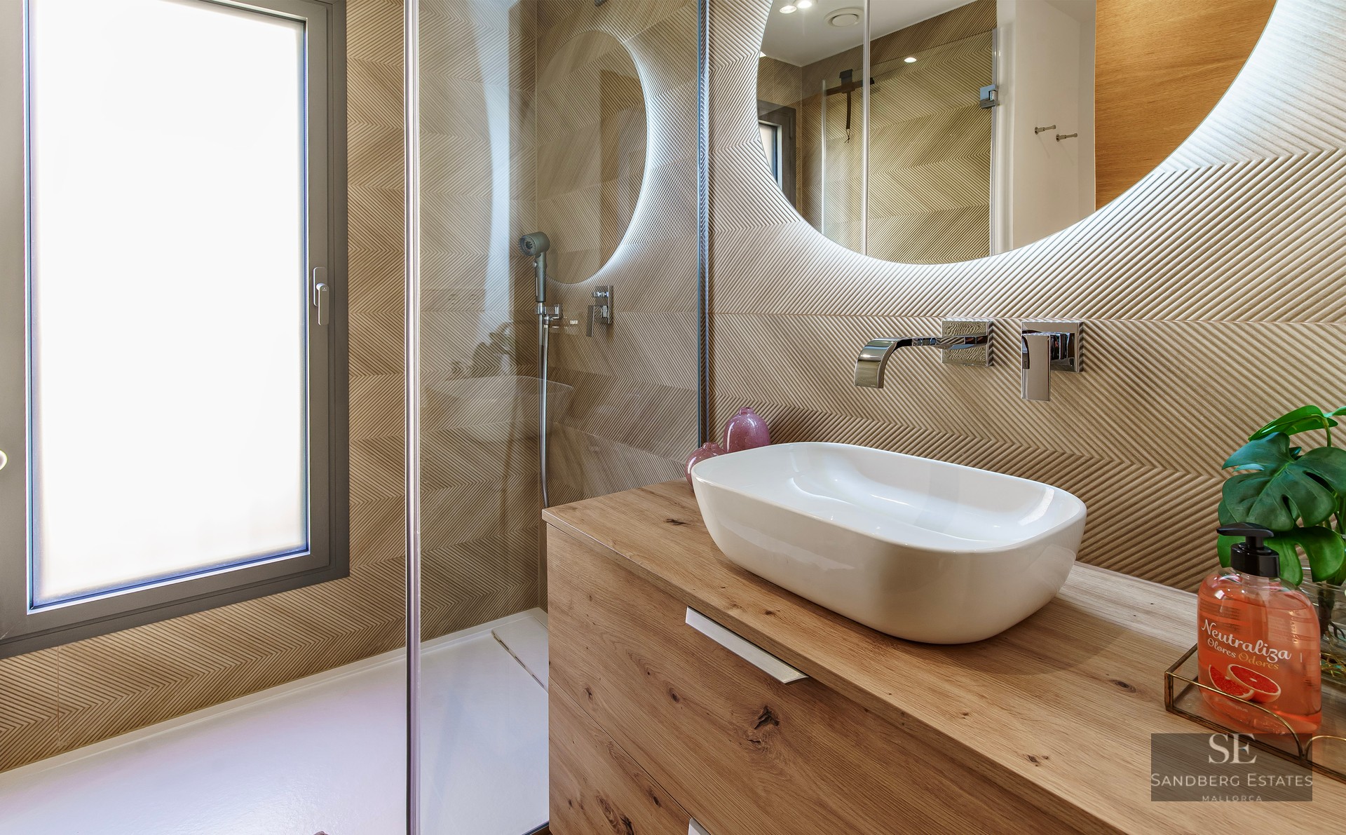 Modern bathroom featuring a wooden vanity, vessel sink, backlit round mirror, and a walk-in shower with glass screen.