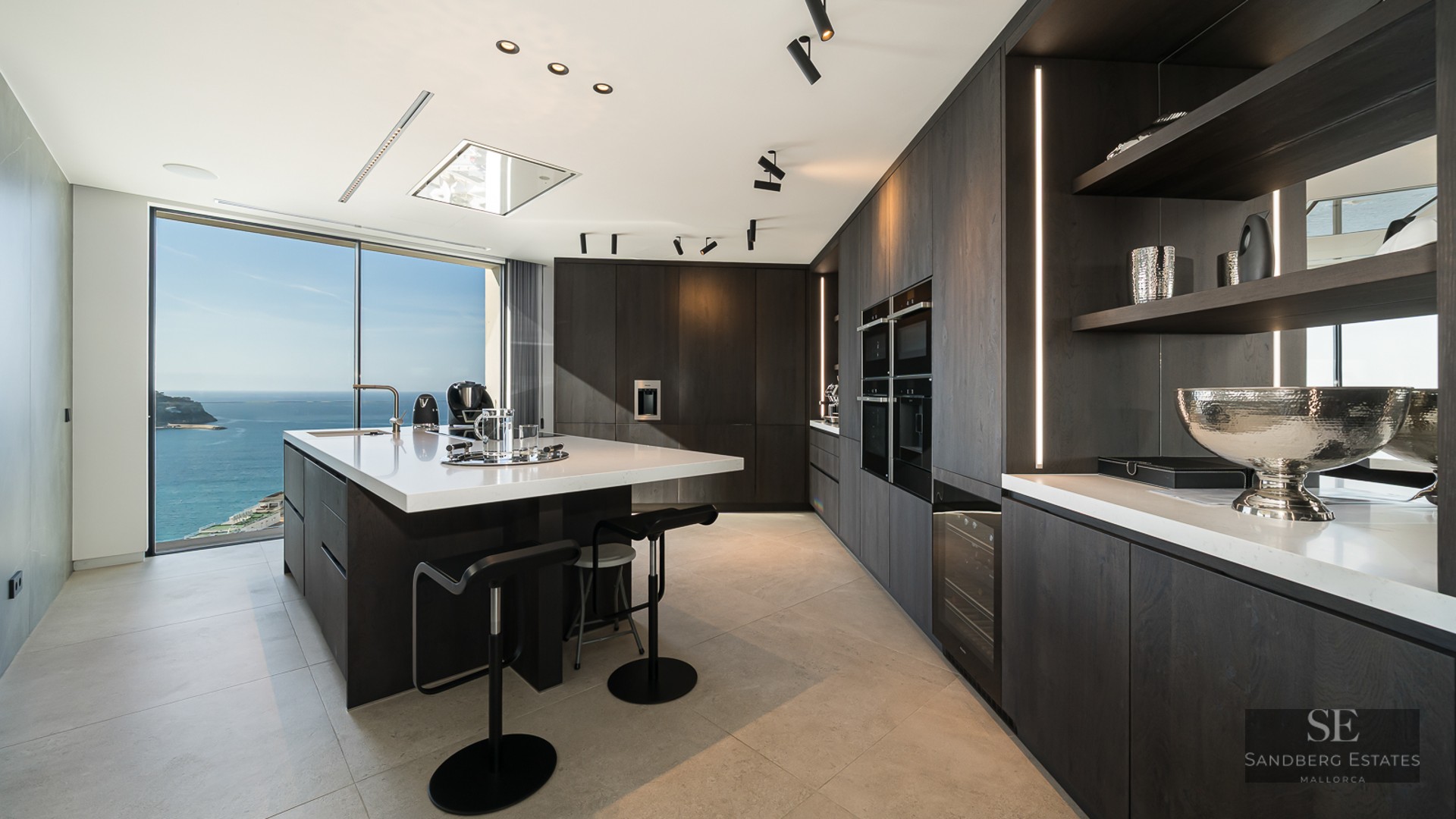 Contemporary dark wood kitchen with a large white island and panoramic ocean views through floor-to-ceiling windows.