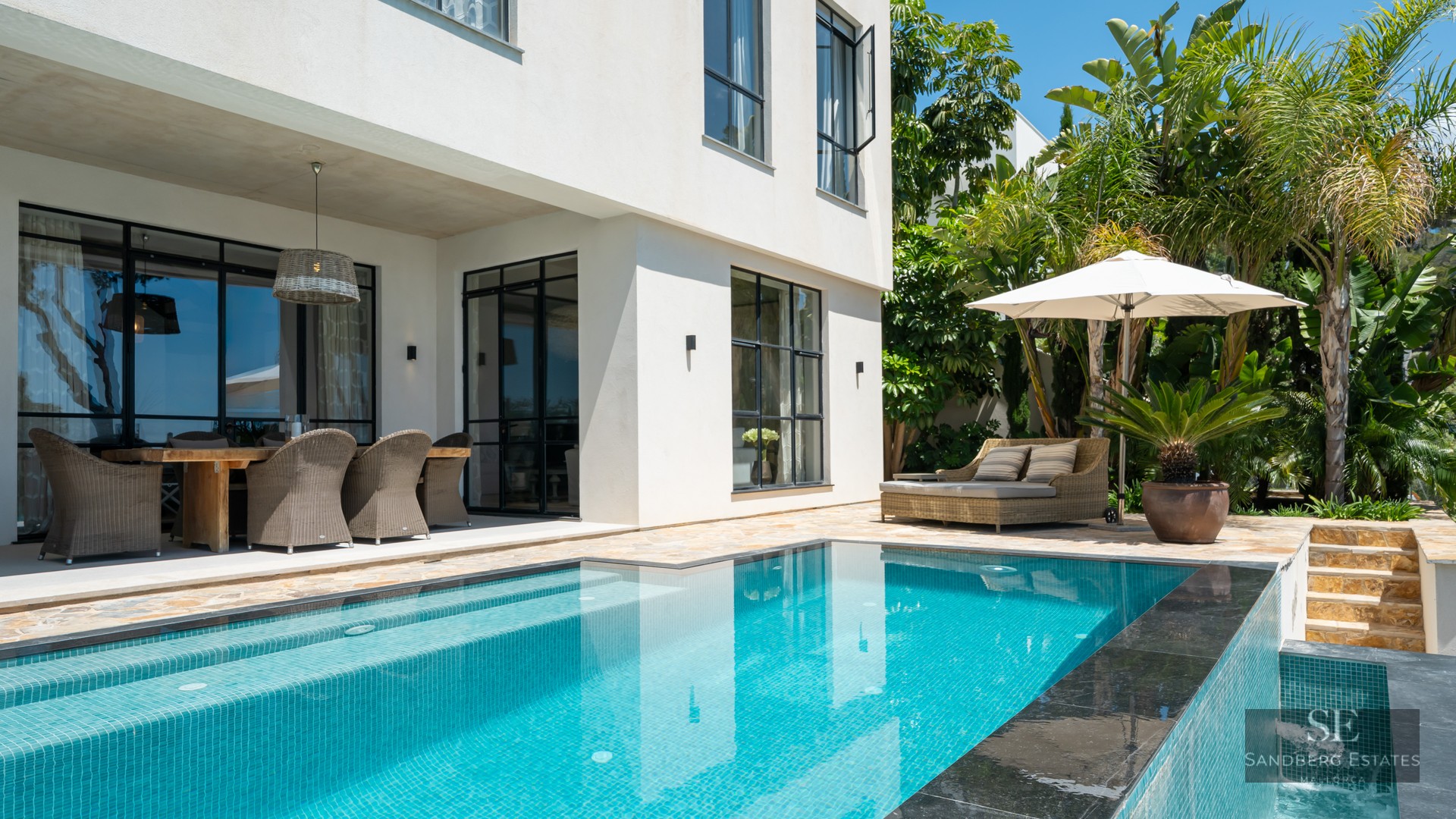Luxury swimming pool next to a white villa with an outdoor dining area, daybed, and tropical garden.