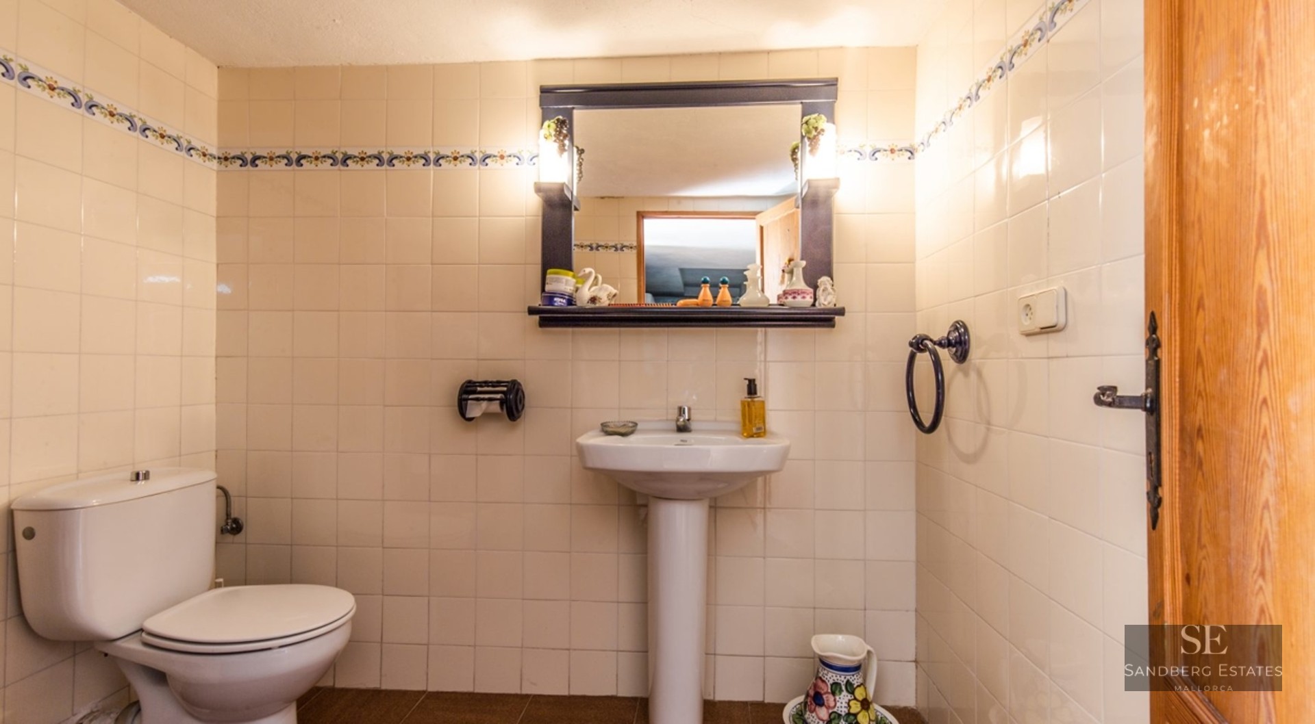Cream tiled bathroom featuring a pedestal sink, toilet, and a dark framed mirror with integrated lighting.