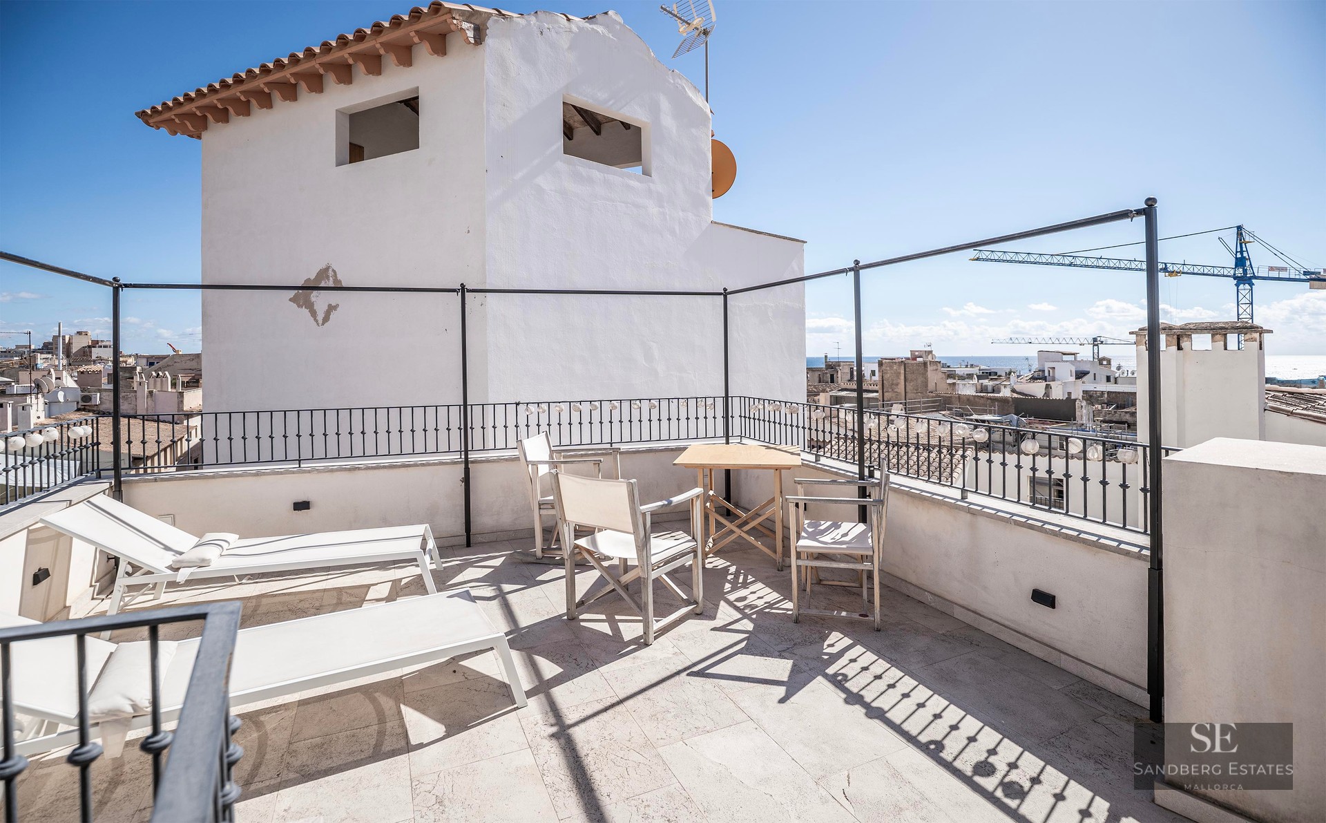 Terrasse ensoleillée avec transats blancs, table en bois et vue lointaine sur la mer au-dessus des toits.